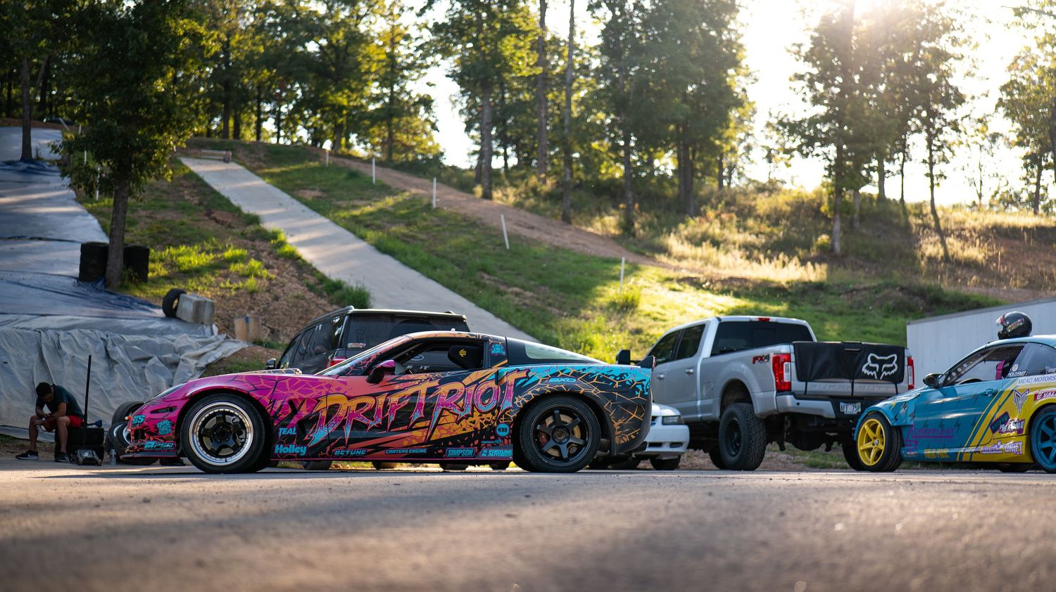 Row of modified cars parked along a tree-lined street during a car meet.