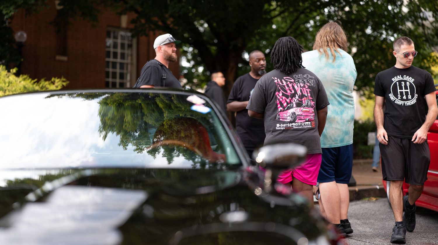 Car enthusiasts gather at a daytime automotive meet in an urban setting.