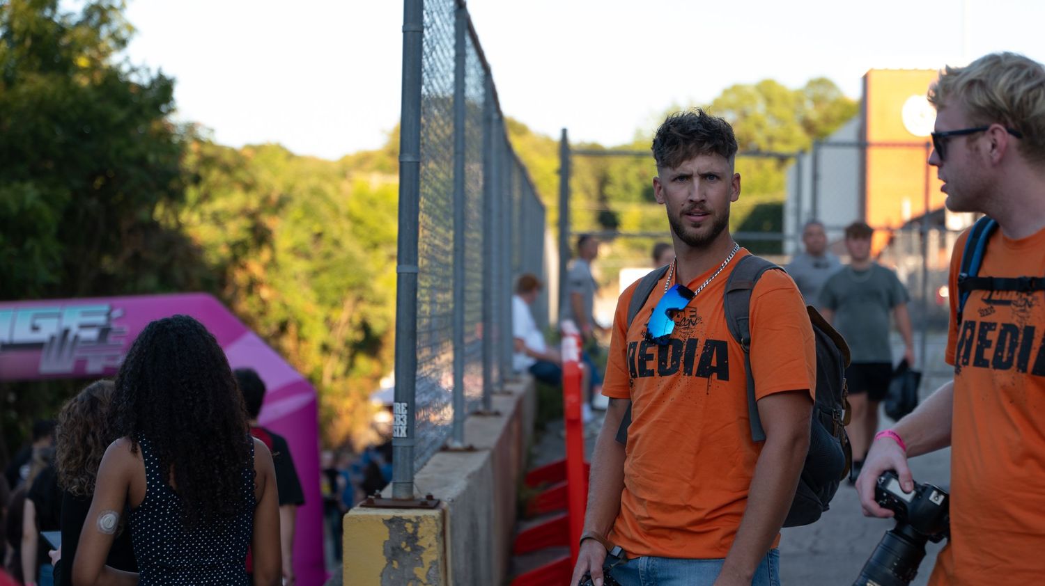 People in orange shirts walking through an outdoor event space with fencing.