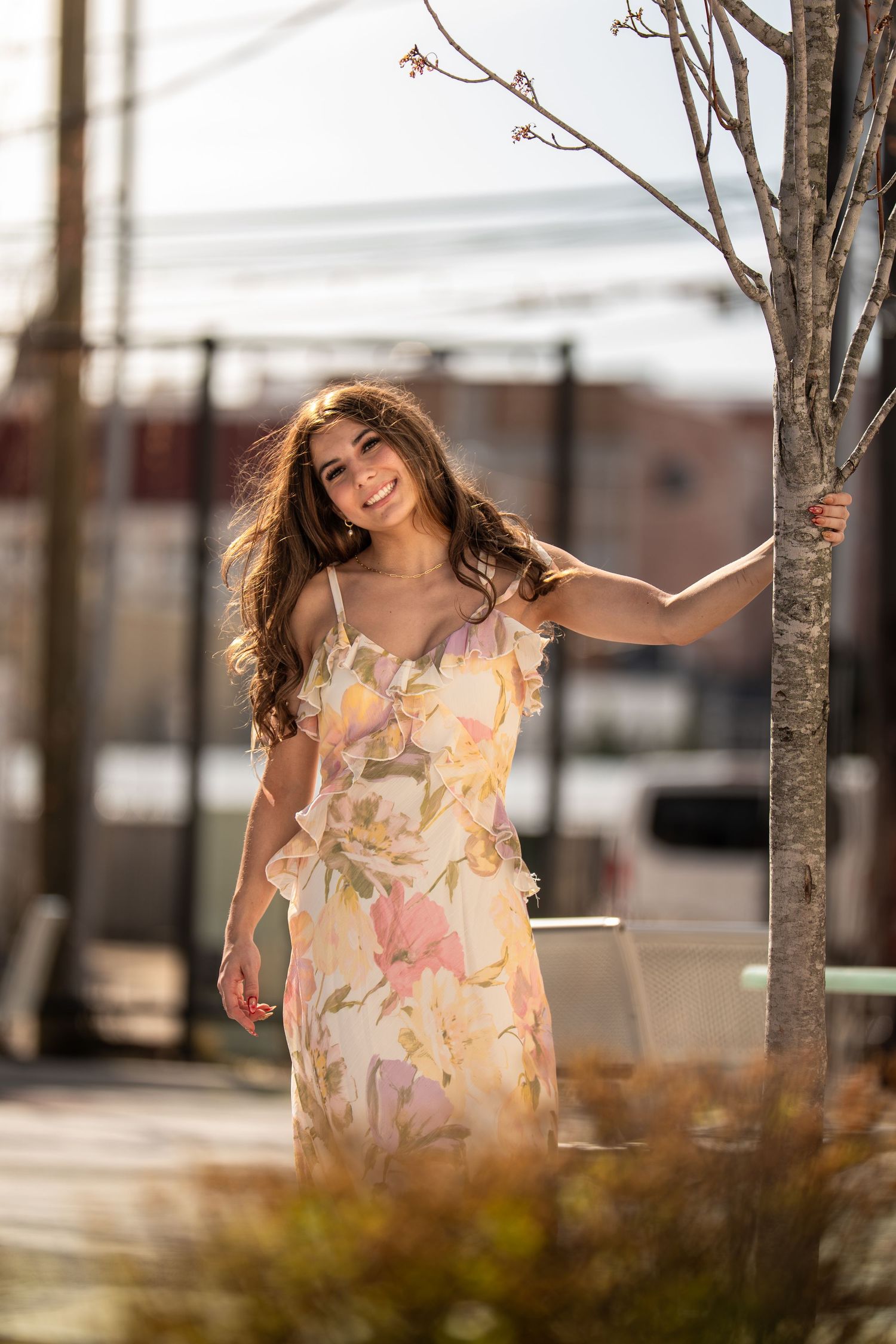 Young woman in floral maxi dress smiling outdoors, leaning against a bare tree in warm sunlight.