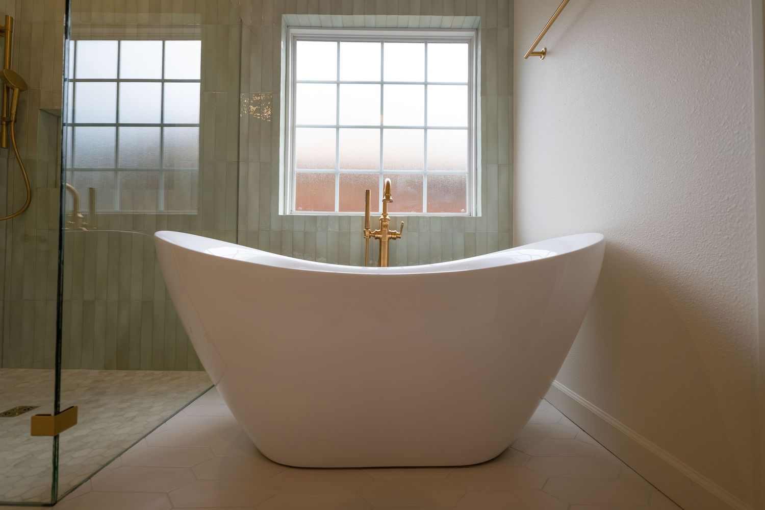 Elegant freestanding white soaking bathtub with gold faucet beside a frosted glass shower and window in a modern bathroom.