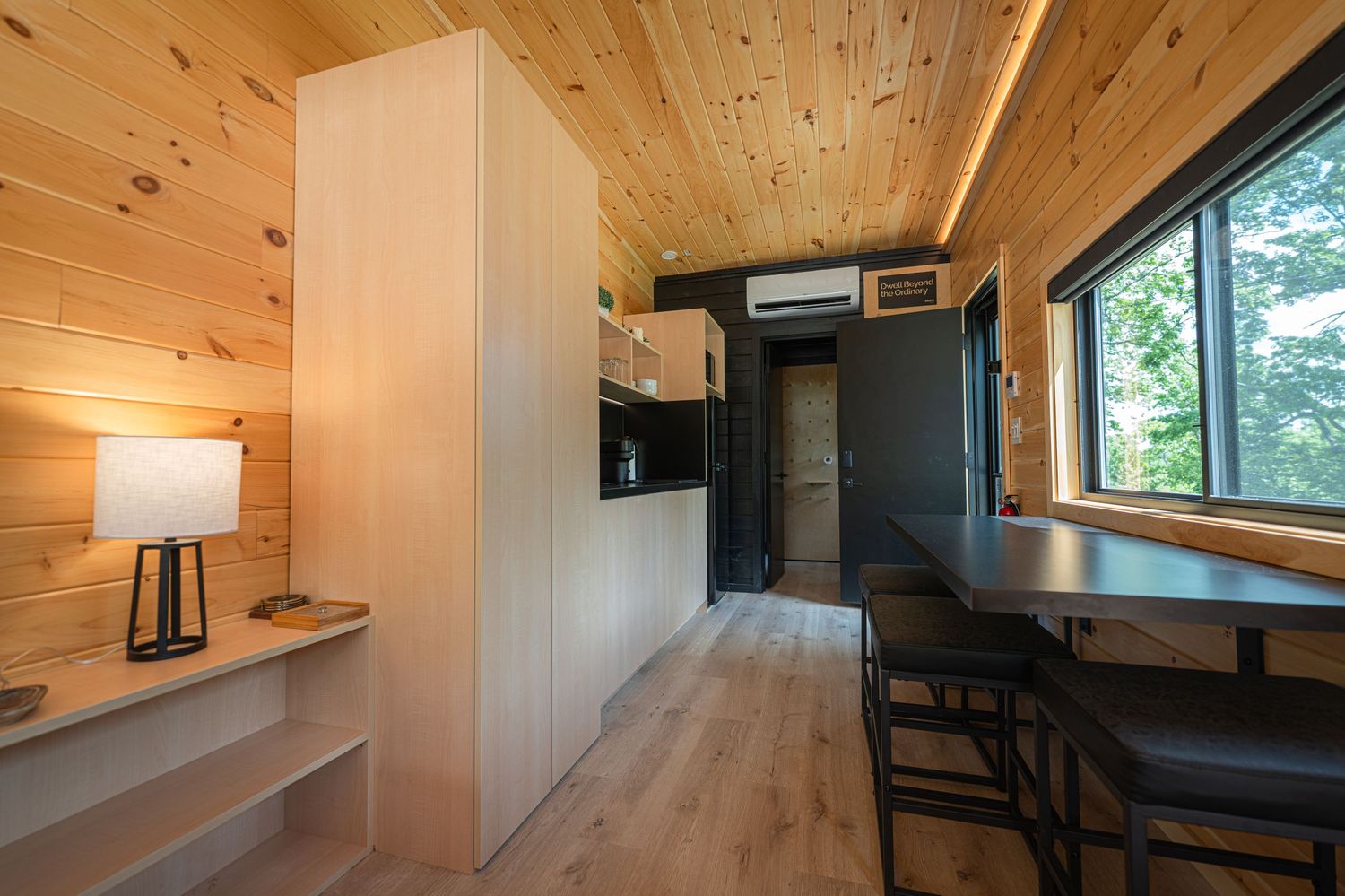 Cozy tiny home interior with wood-paneled walls, light hardwood floors, mini kitchen, dining table, and large window overlooking trees.