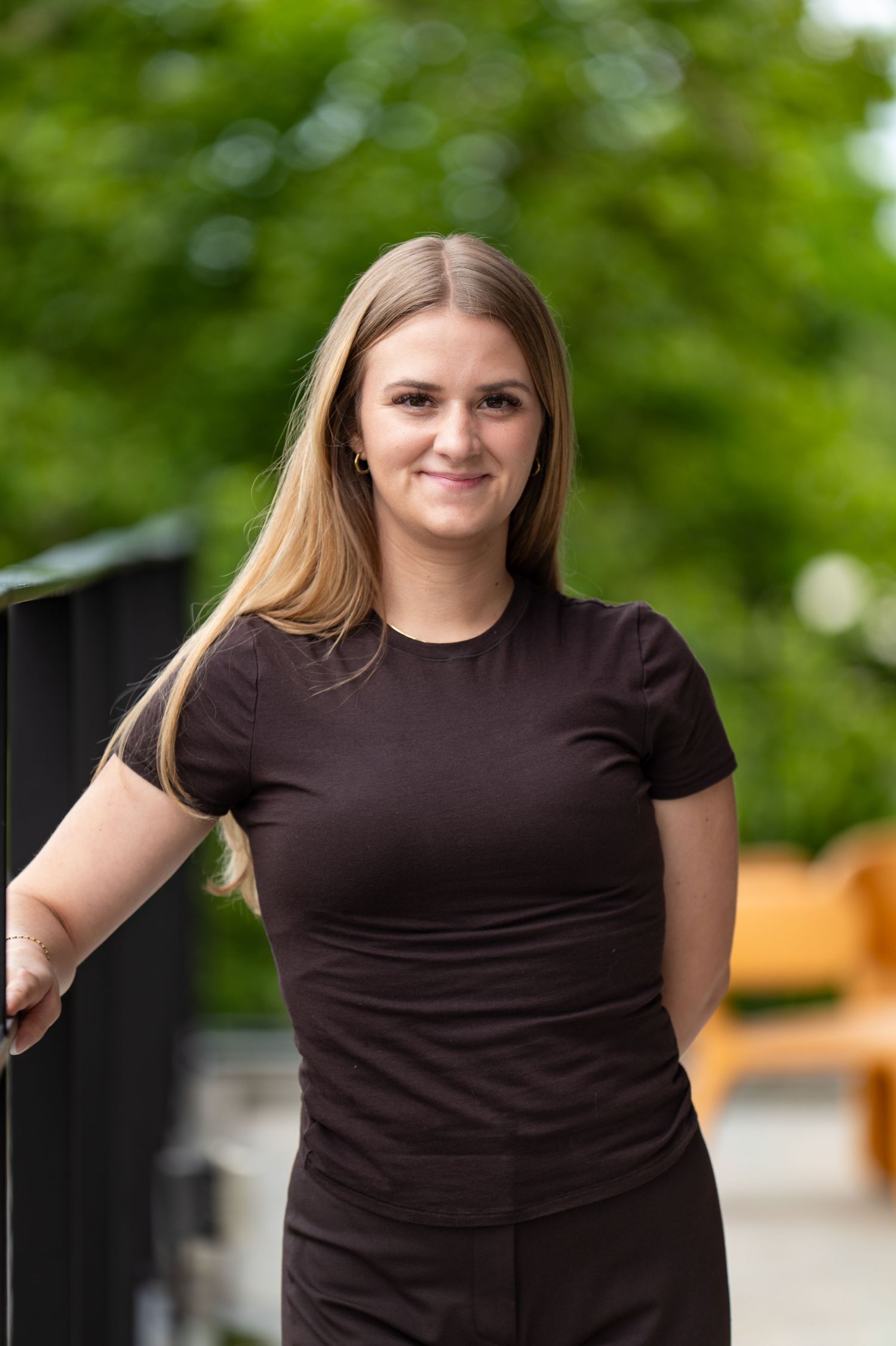 Young woman in black outfit smiling outdoors with lush green trees in background.