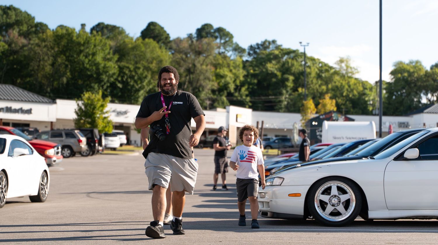 People walking through parking lot with modified cars at automotive event.