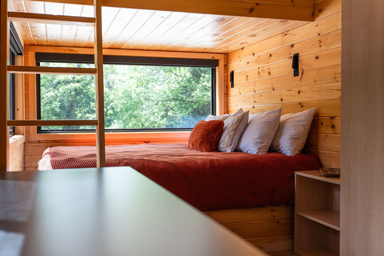 Cozy wooden cabin bedroom with a red bedspread, white pillows, and a large window overlooking lush green trees.