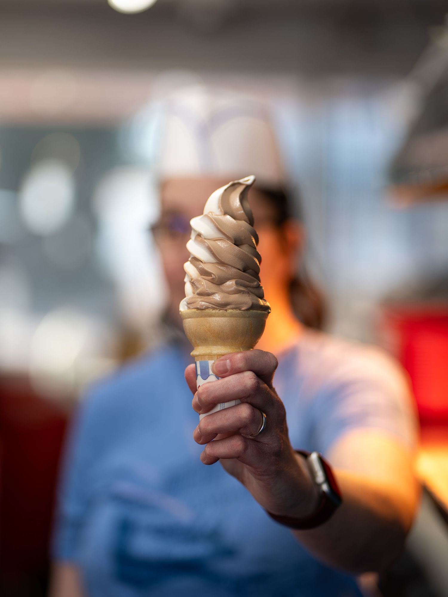 A hand holds up a swirled soft-serve ice cream cone against a blurred background.