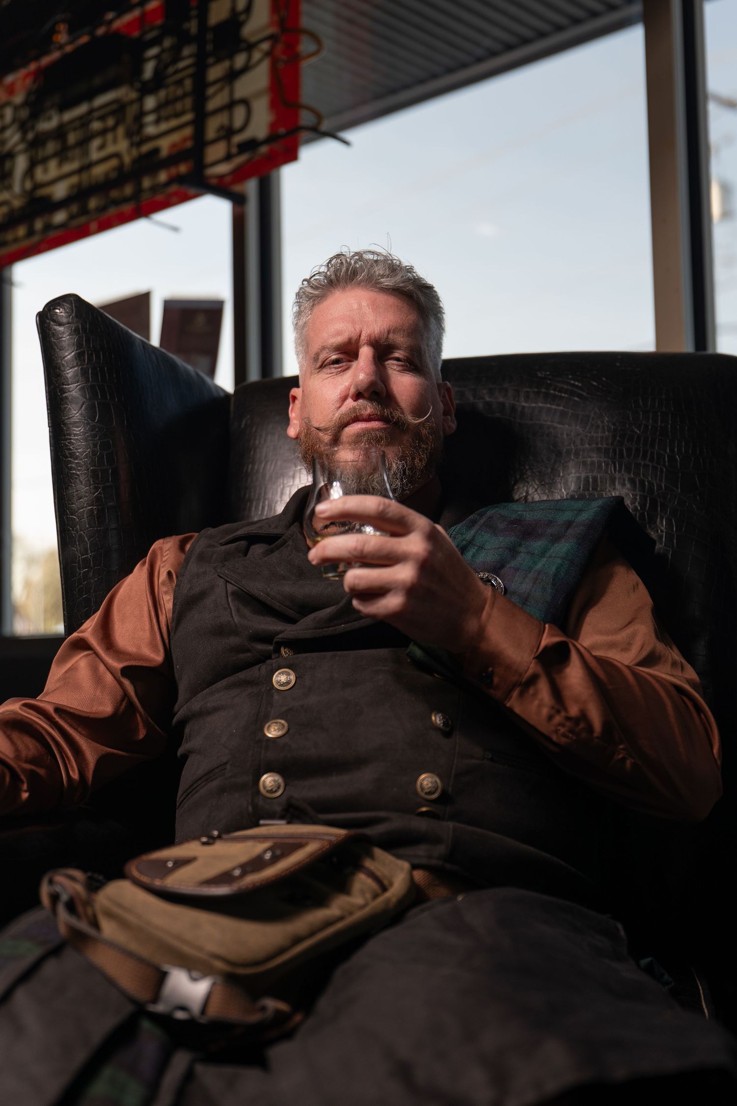 Bearded middle-aged man in vest holding whiskey glass, relaxing in leather chair near window.