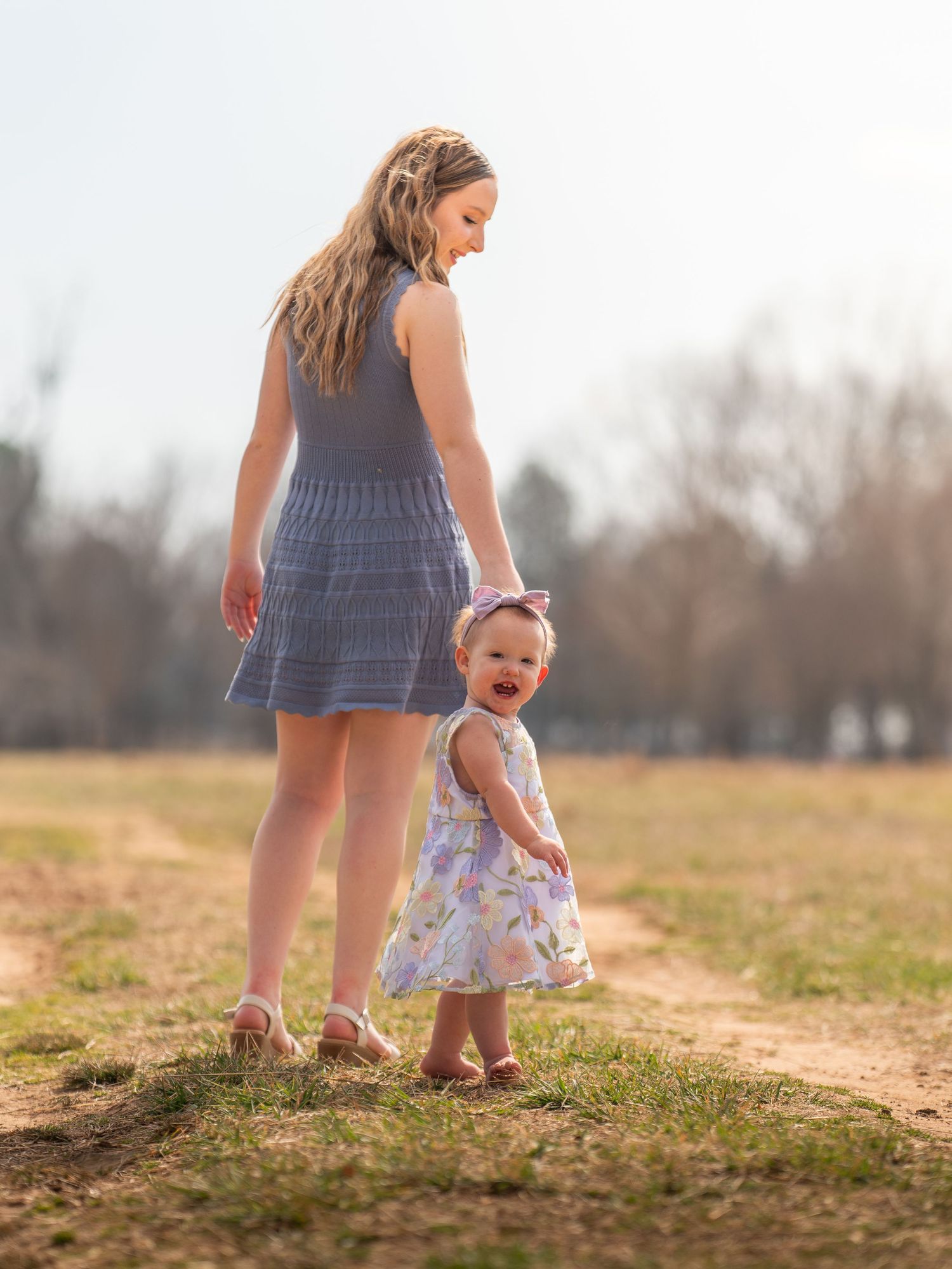 A mother in a blue dress holds her laughing baby girl in a floral dress while walking on a sunny park path.
