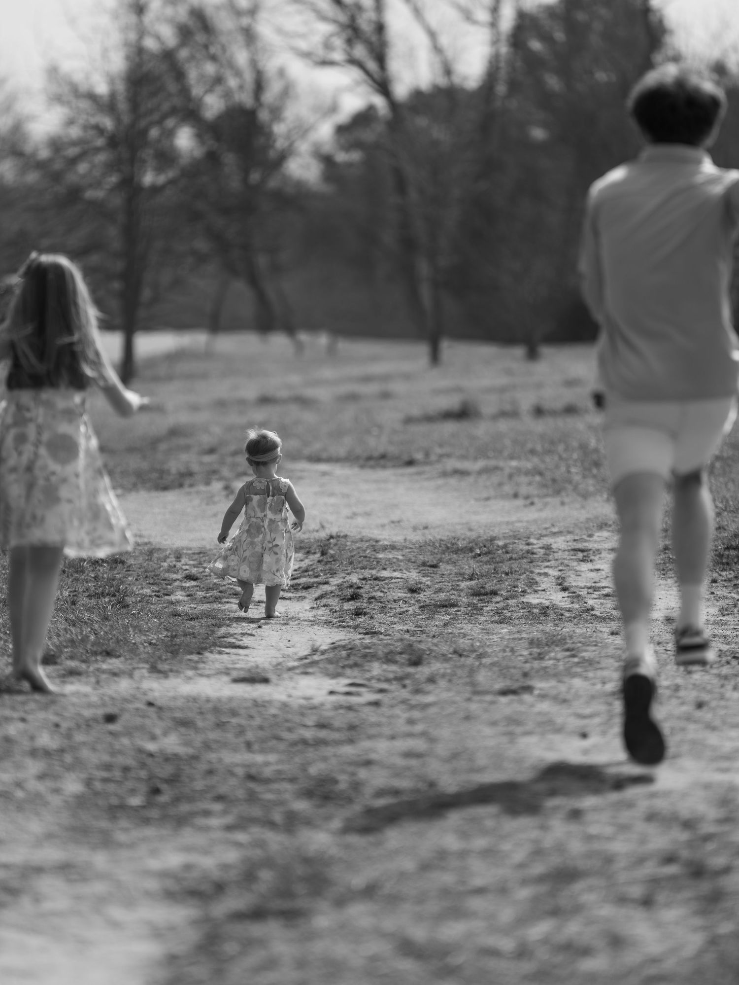 Black and white photo of three children running on a dirt path in a park, with trees in the background.
