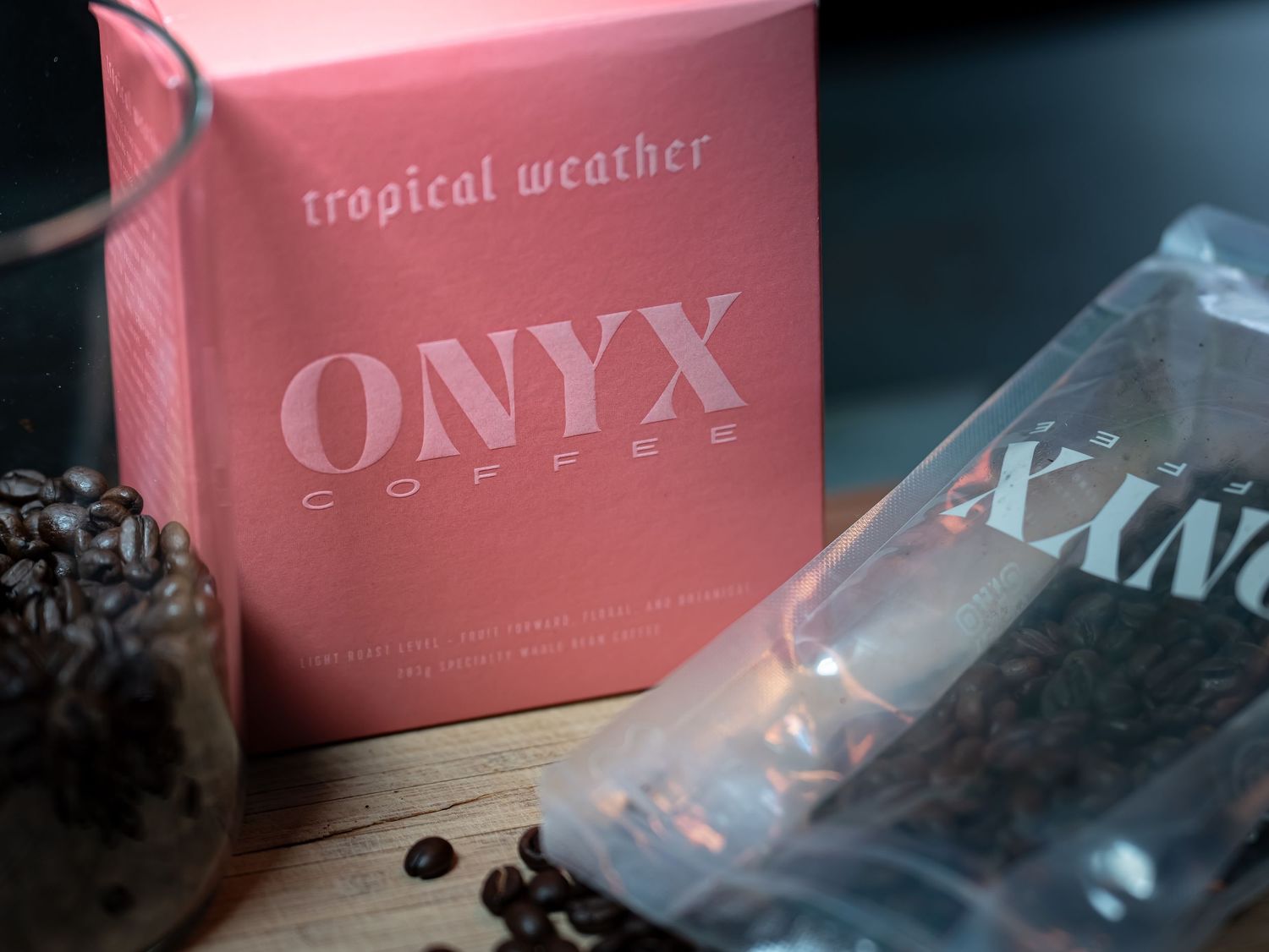 Pink Onyx Coffee Tropical Weather blend box with coffee beans and clear packaging on a dark wooden surface.