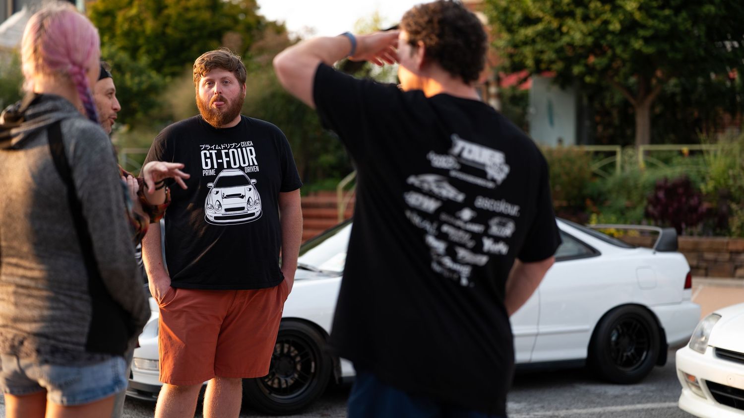 People socializing at a car meet during golden hour with vehicles parked in background.