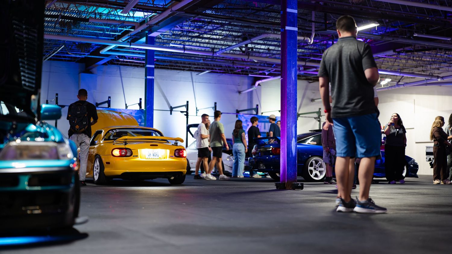 Car show in dimly lit garage space with blue LED lighting and crowds gathering around vehicles.