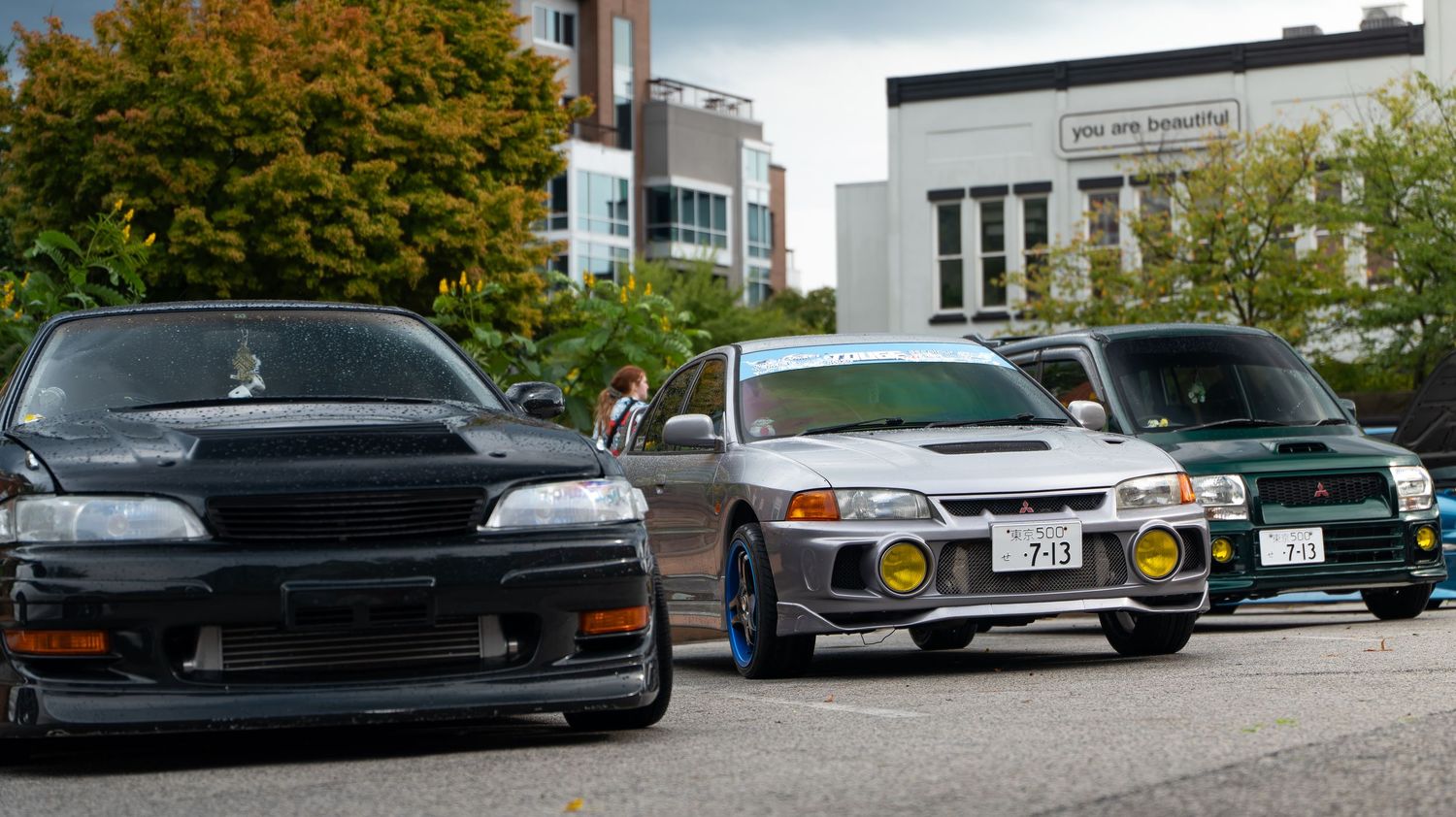 Line of modified sports cars displayed at an automotive event with fall foliage in background.