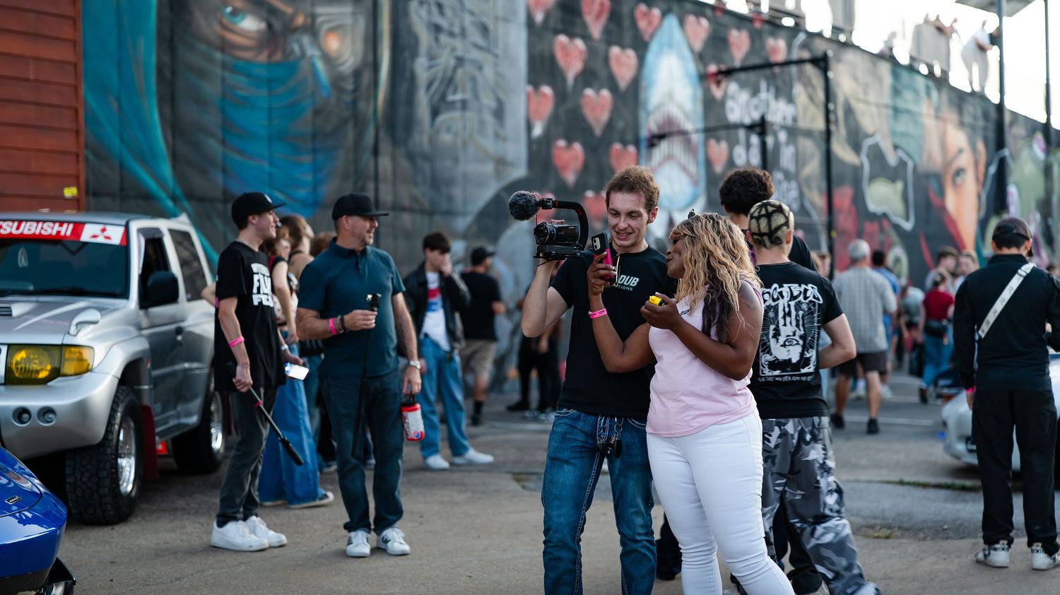Long panoramic view of a car meet with crowds gathering along a wall decorated with murals.