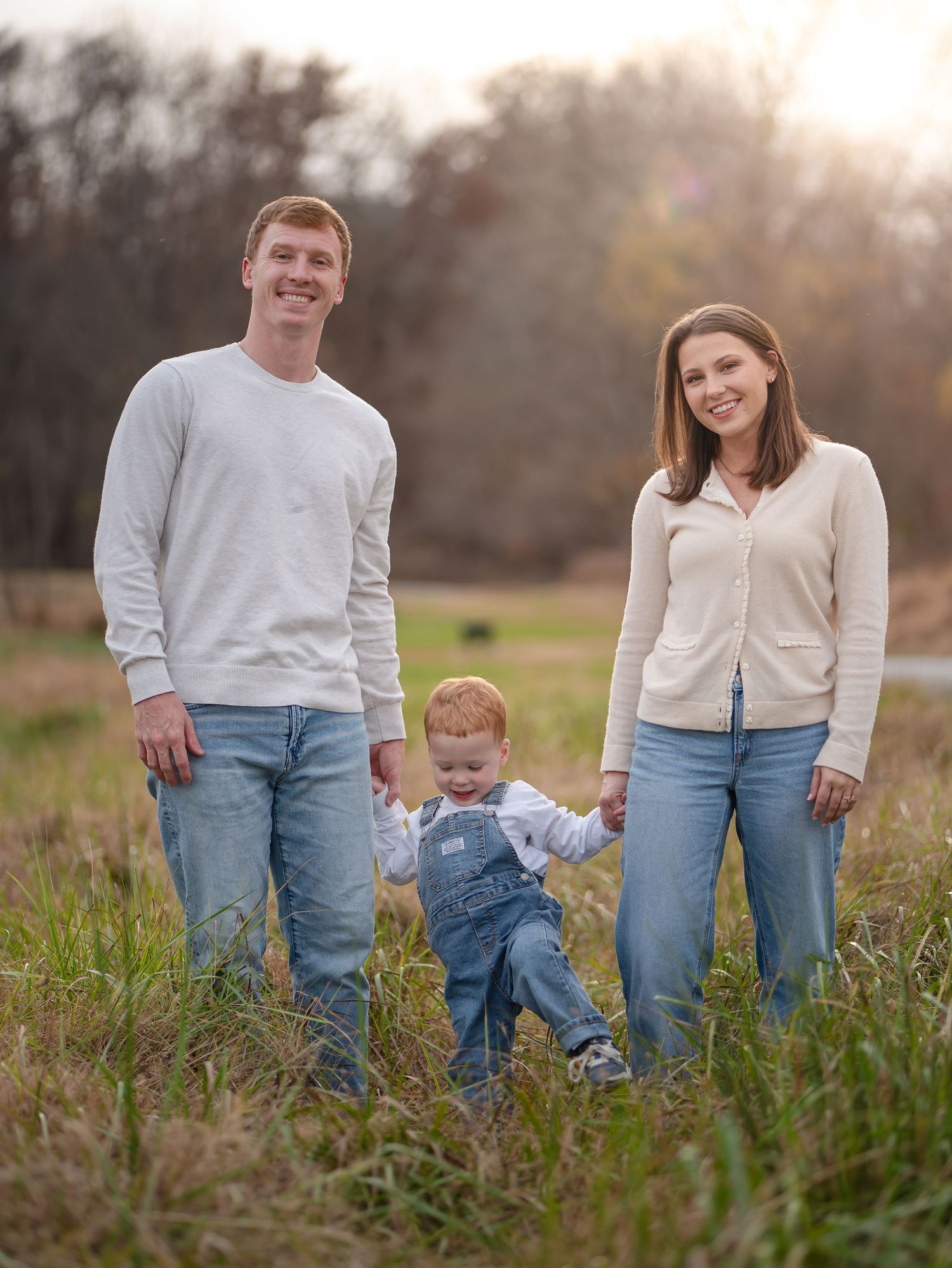 A family of three poses together in a grassy field during golden hour wearing casual clothing and denim.