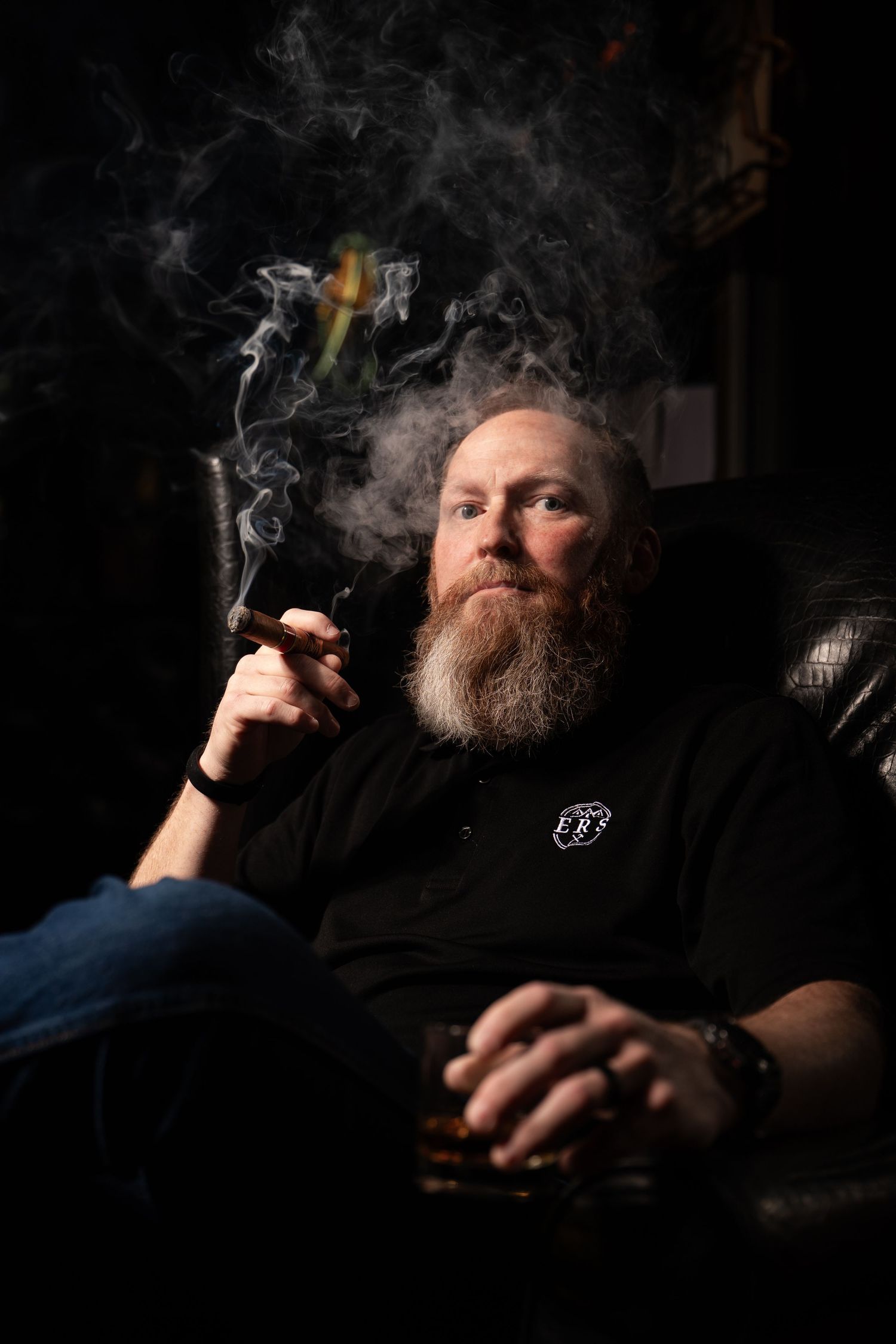 Bearded man smoking a cigar and holding whiskey, relaxing in a leather chair in a dark room.