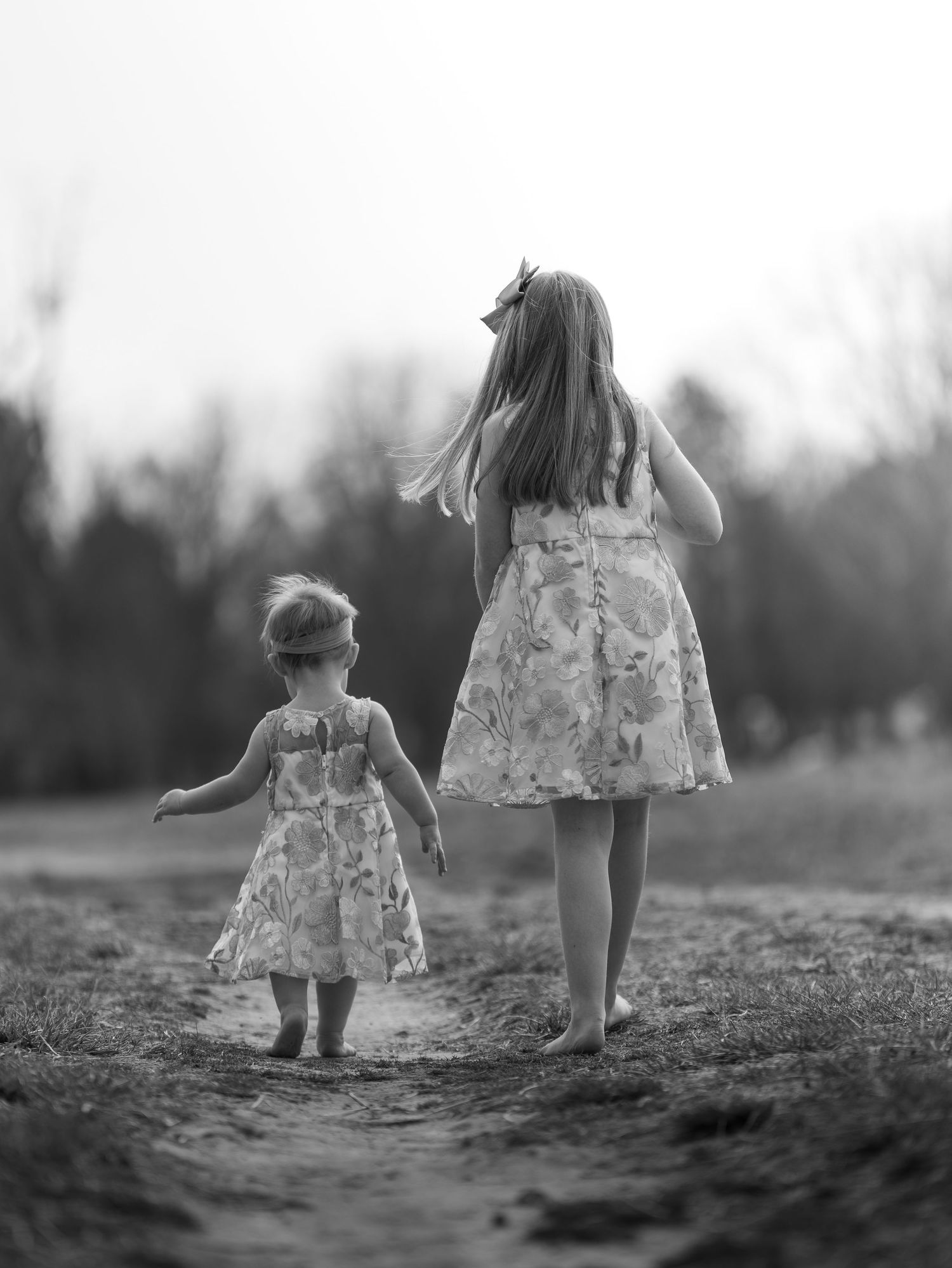 Two young girls in matching floral dresses walk hand in hand down a dirt path in a black and white photo.