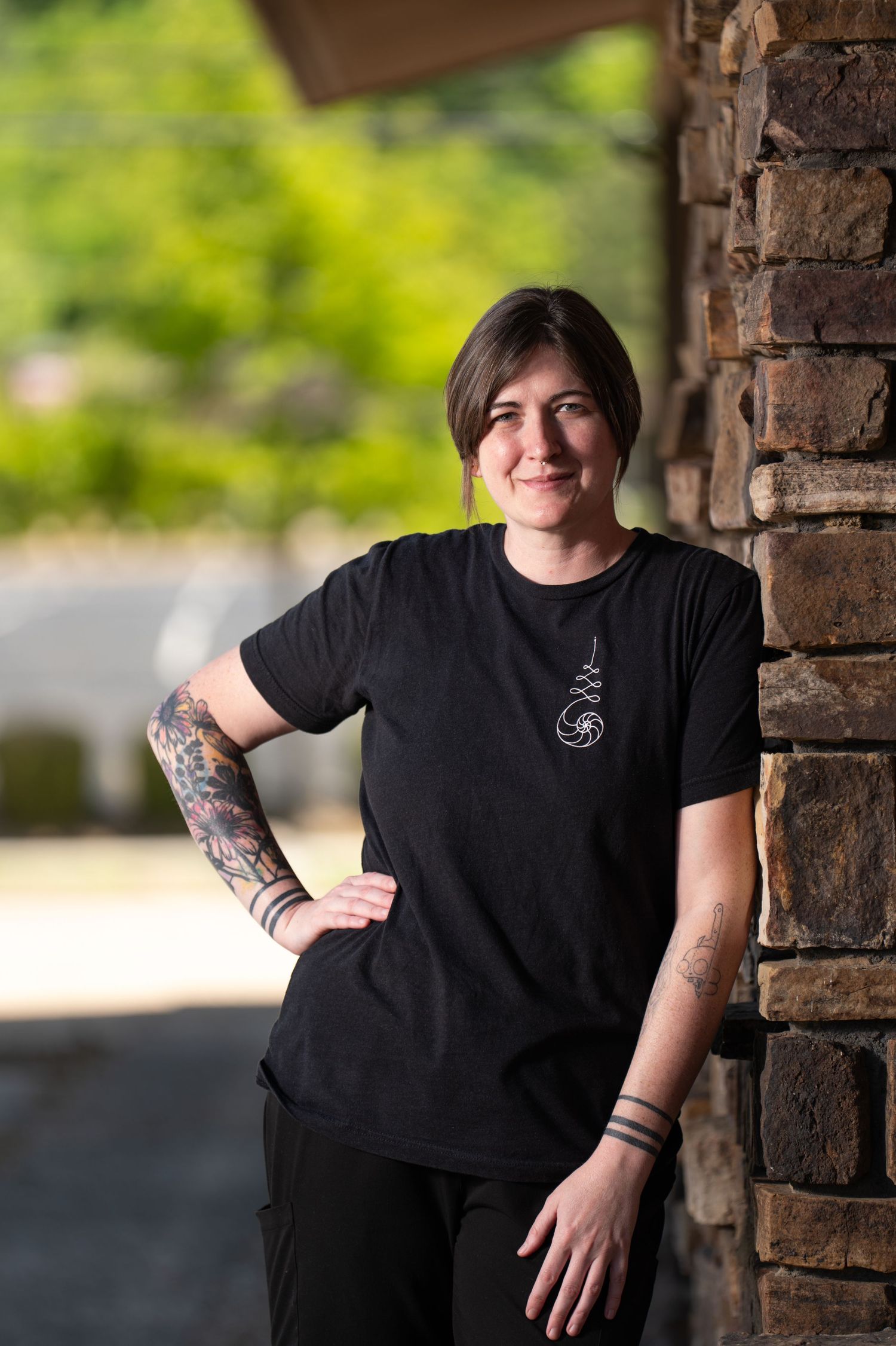 Smiling woman with tattoos in black t-shirt leans against stone wall outdoors with green trees in background.