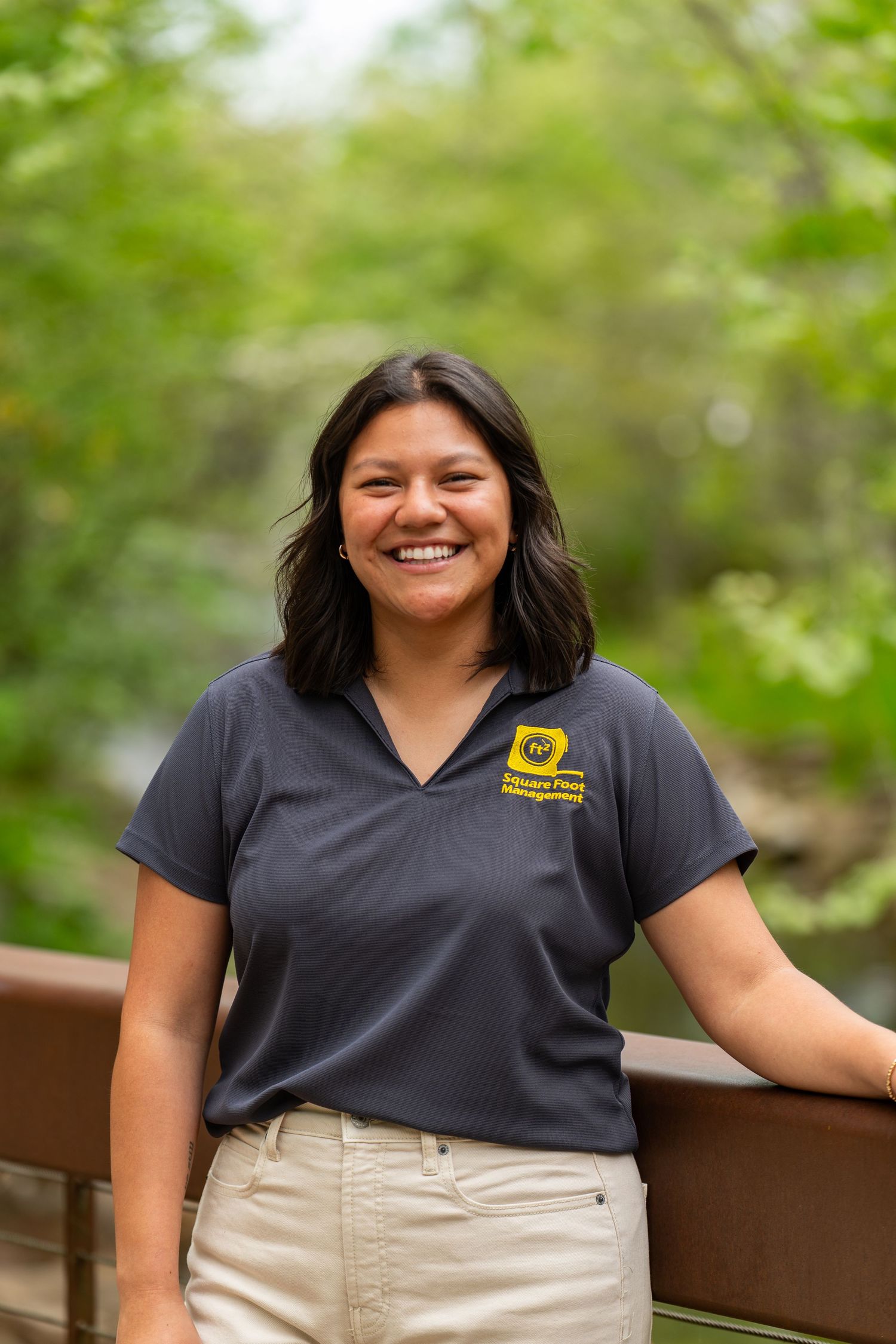 Smiling young woman in navy polo shirt standing on wooden bridge surrounded by lush green trees outdoors.