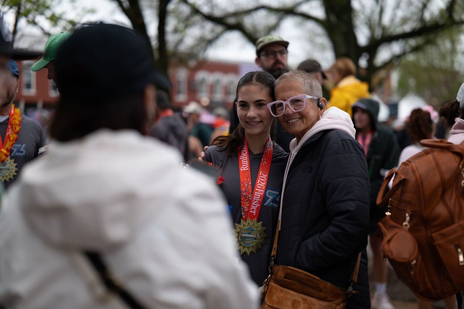 Two people pose for a photo outdoors after a race, one wearing a finisher medal, surrounded by a crowd.