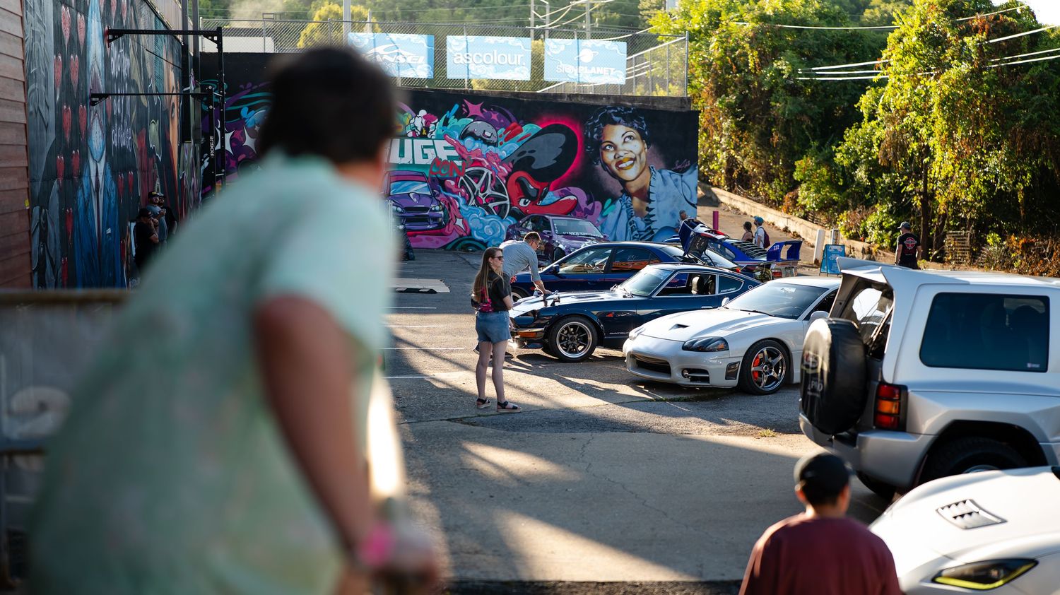 Panoramic view of a car meet showing various vehicles against a backdrop of trees and graffiti.
