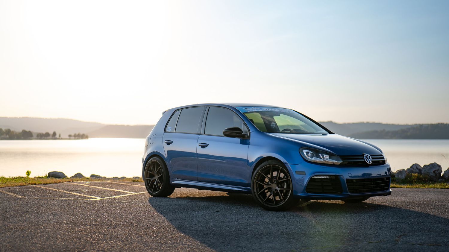Row of modified Volkswagen Golf GTI and R cars lined up at a car meet against a scenic backdrop.