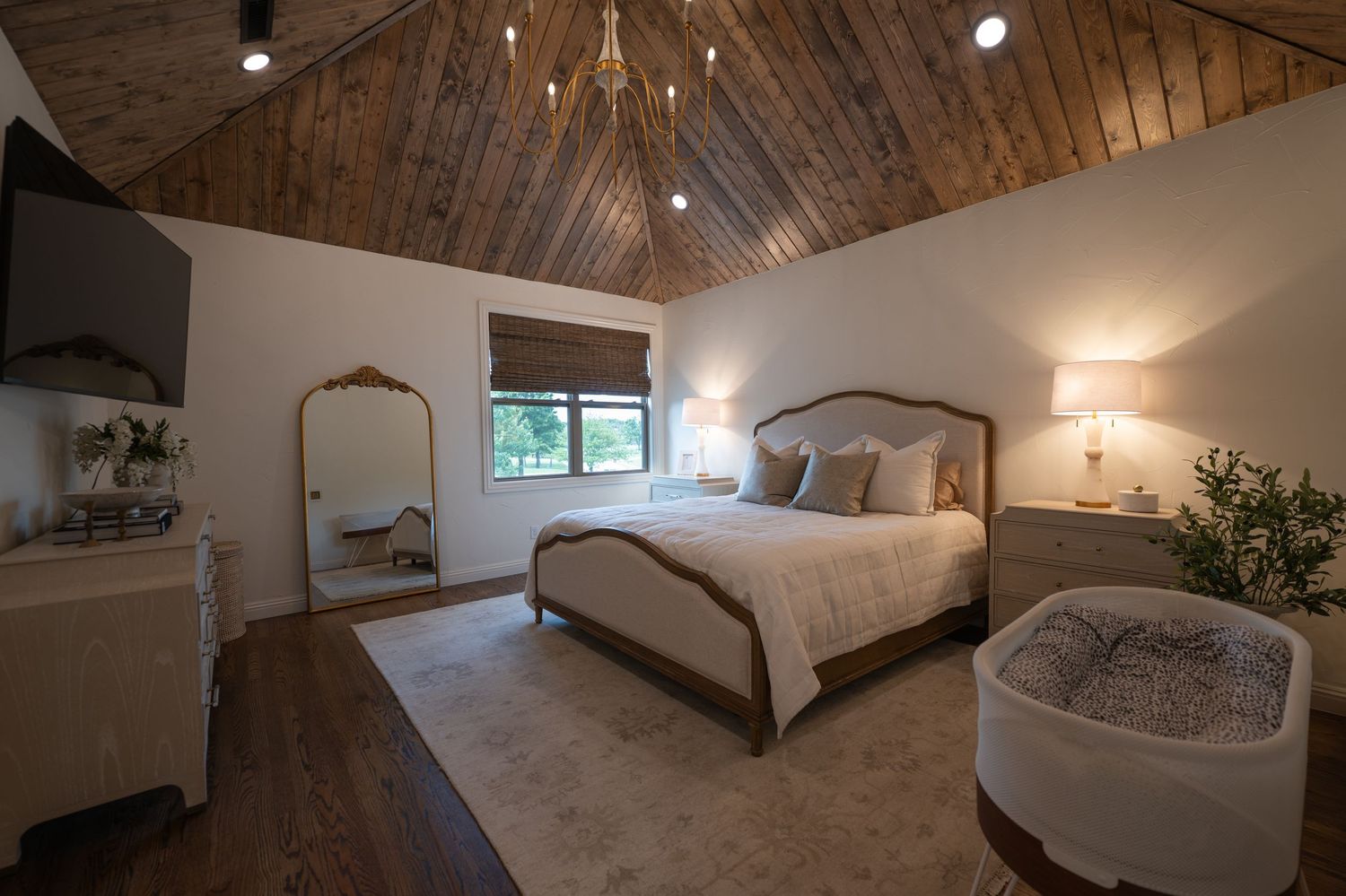Elegant bedroom with vaulted wood-plank ceiling, chandelier, upholstered bed, hardwood floors, and warm neutral decor.
