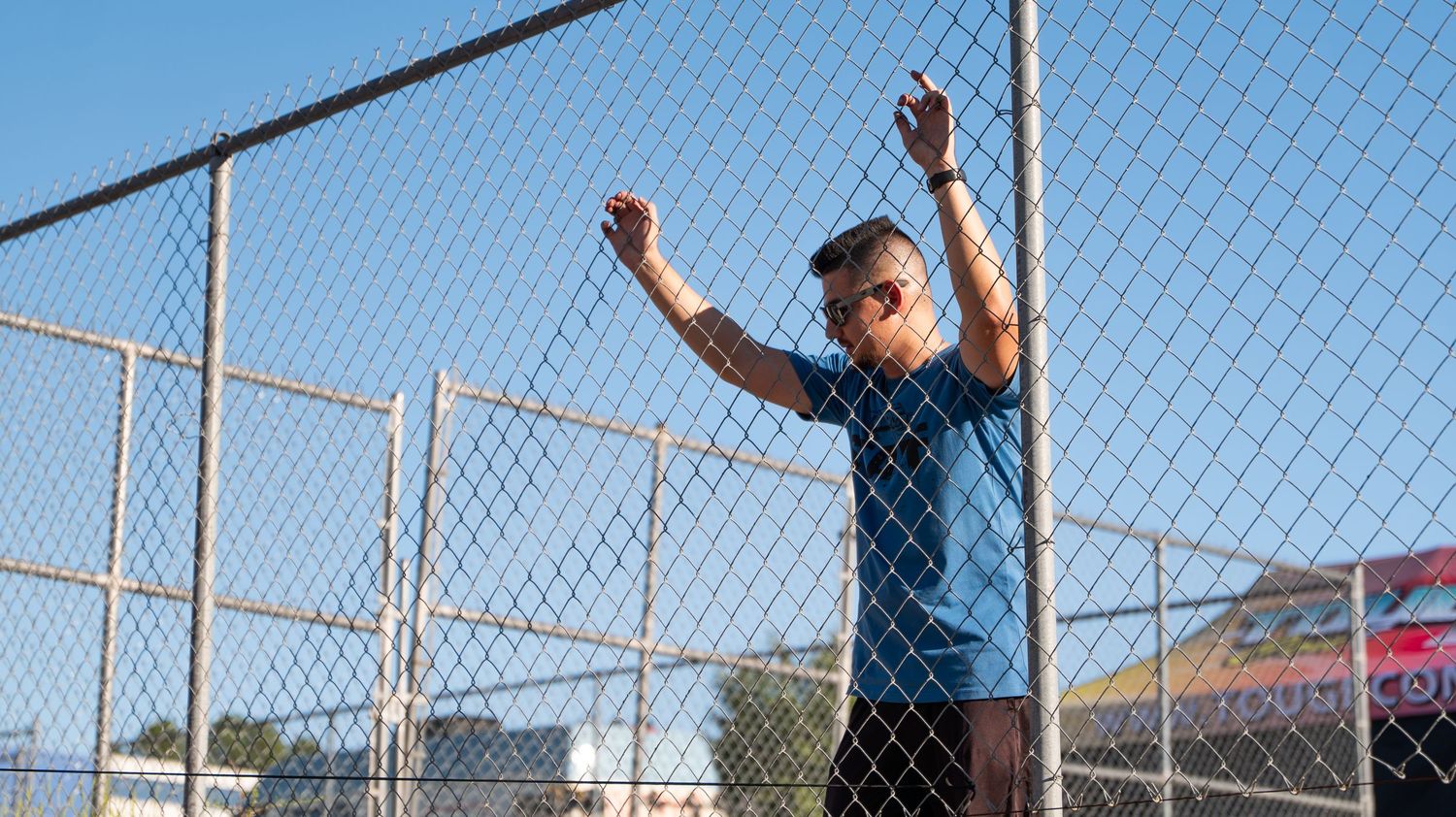 Person in blue shirt poses against chain link fence at outdoor event.