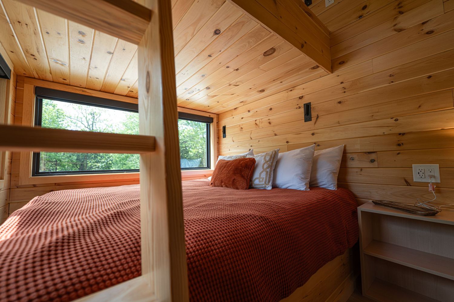 Cozy wooden cabin bedroom with red plaid bedding, bunk bed, and large windows overlooking lush green trees.