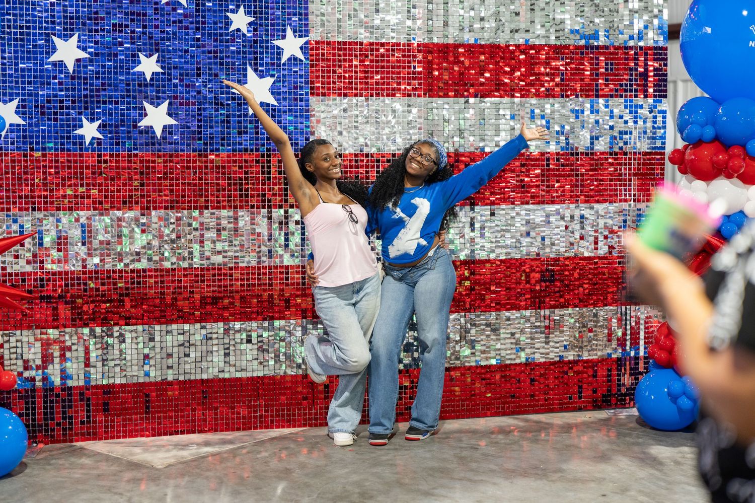 Two women pose joyfully in front of a sequined American flag backdrop with red, white, and blue balloon decorations.