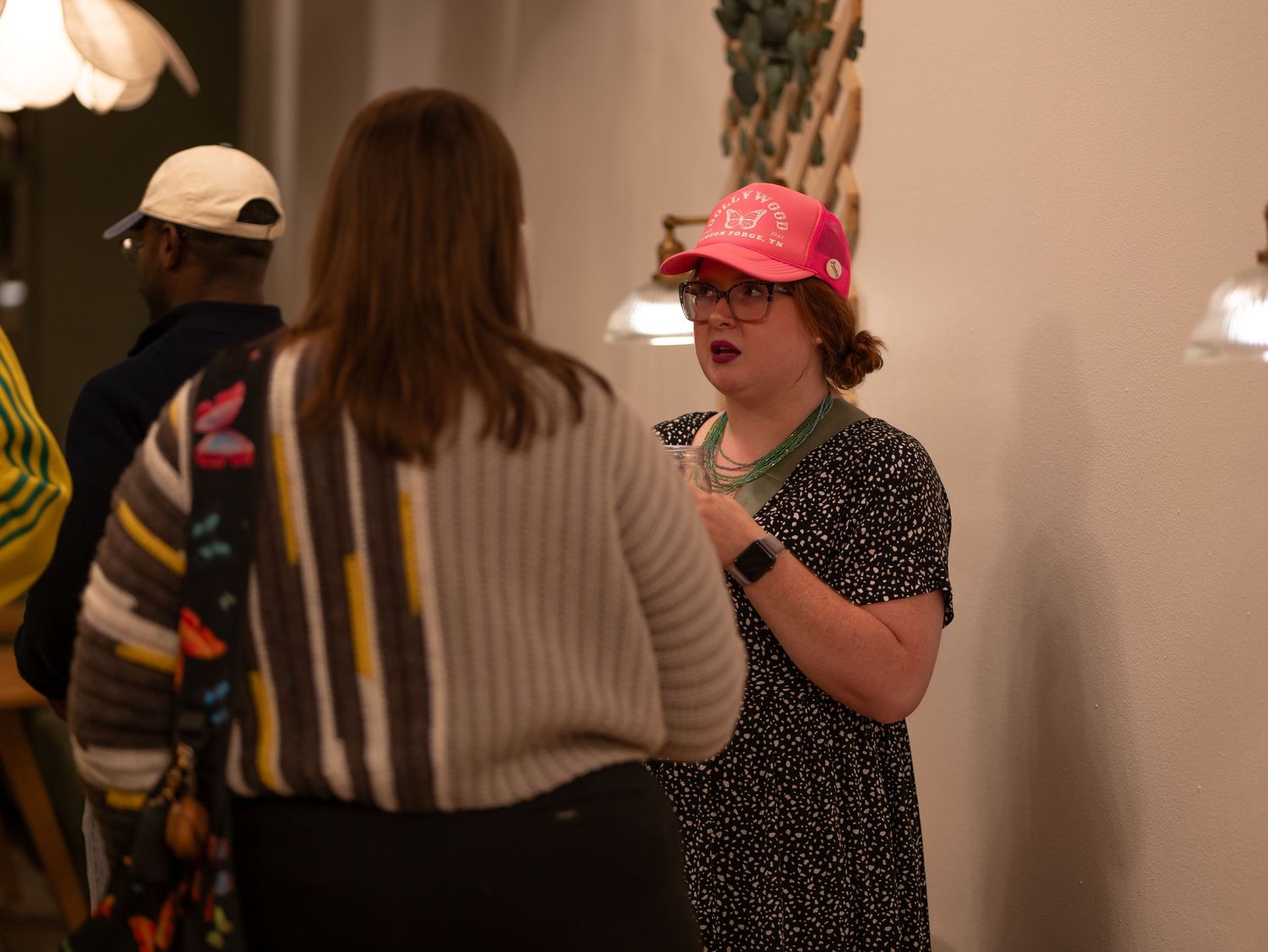 A group of people gather indoors for a social event, with some wearing casual attire and baseball caps.
