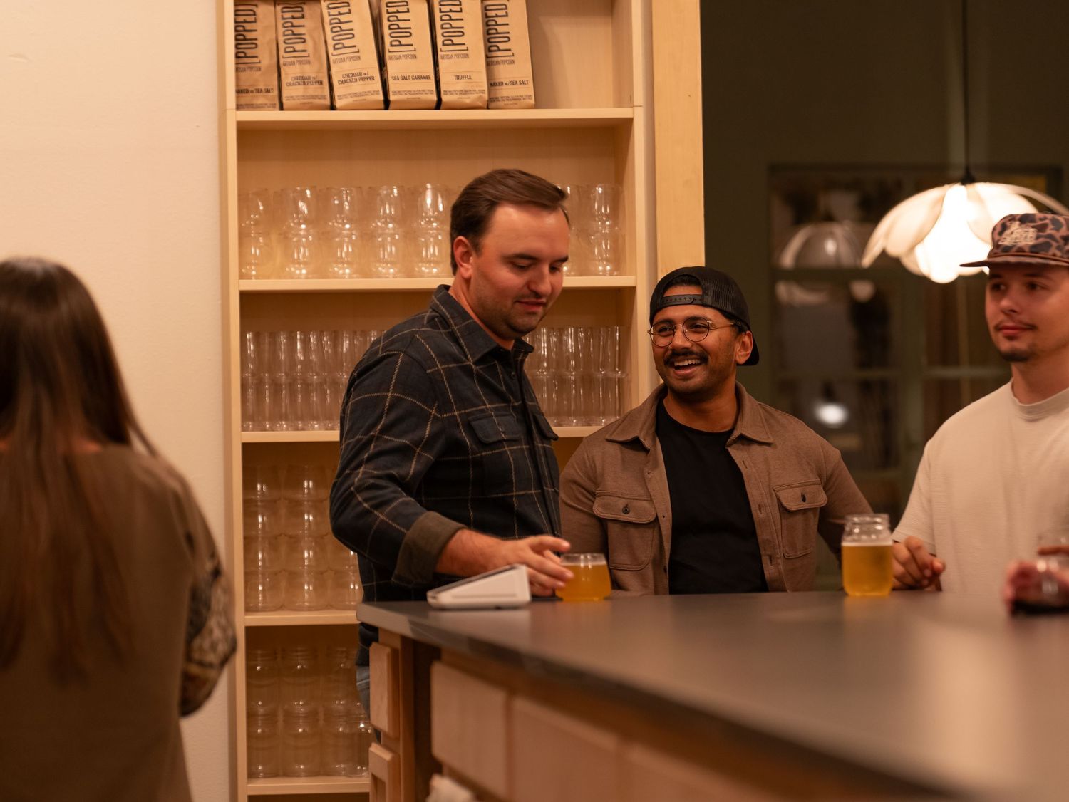 People gathered at a bar counter with wooden shelves of glassware in the background during evening hours.