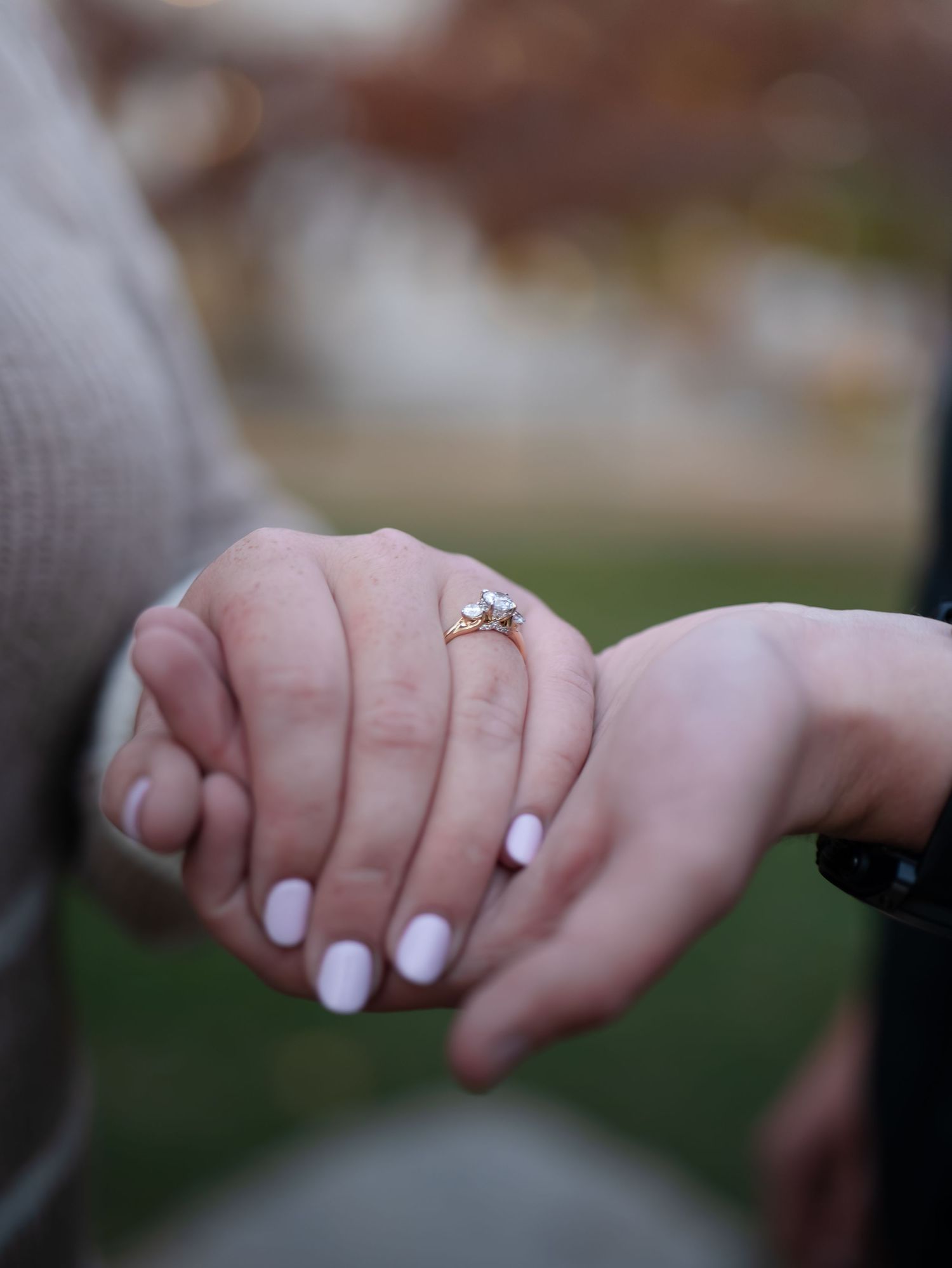 Close-up of hands showing off an engagement ring with white nail polish against a blurred outdoor background.