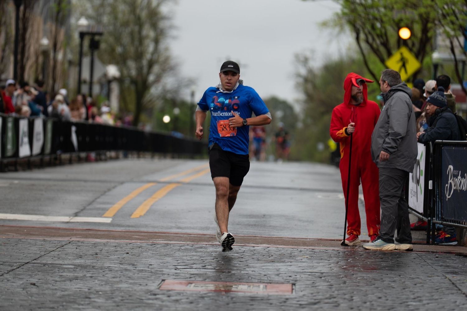 Runner crosses wet finish line in blue shirt at rainy outdoor marathon, spectator in red costume watches nearby.