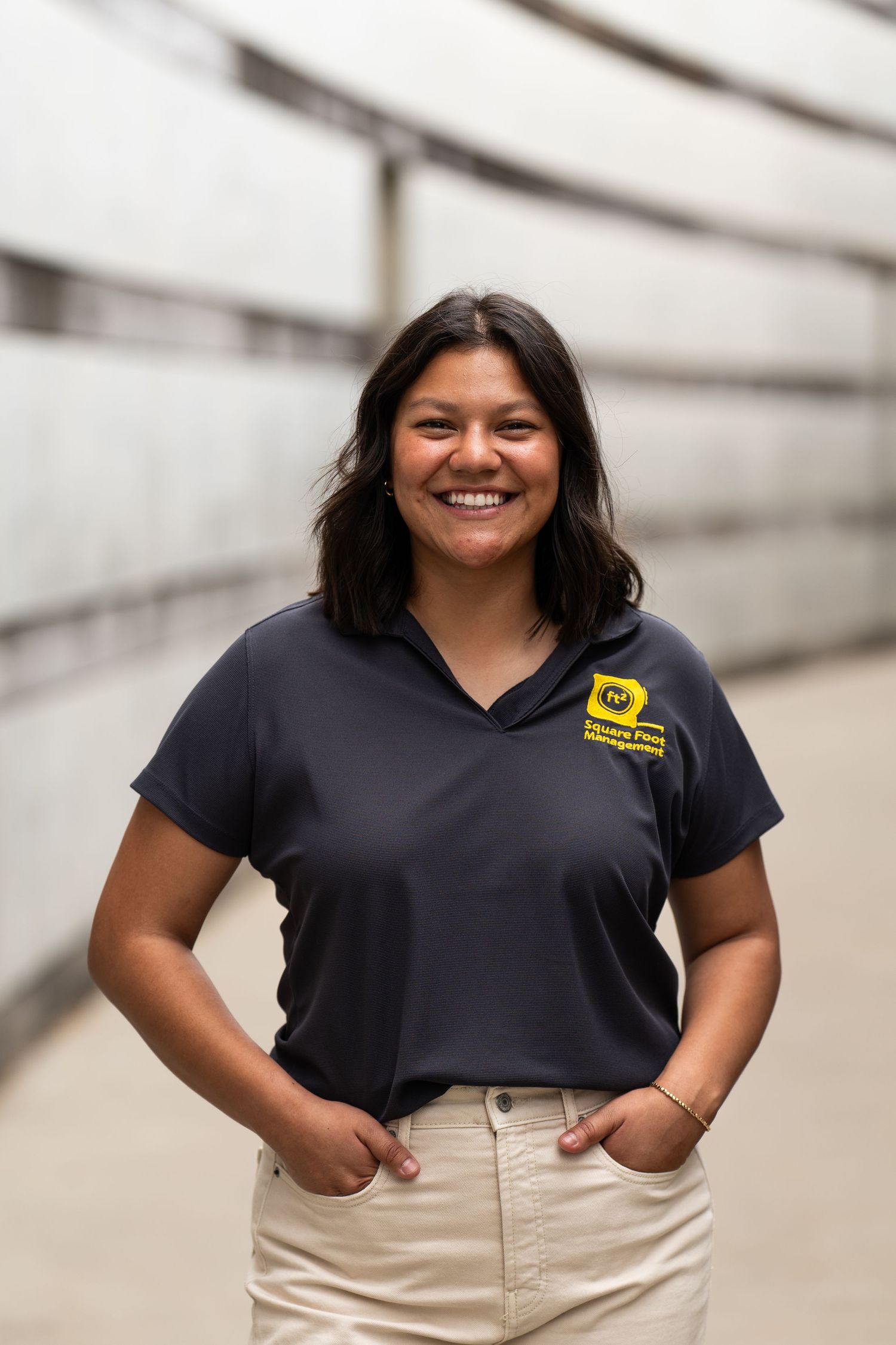 Smiling young woman in a dark navy polo shirt with a yellow logo, standing confidently with hands in pockets outdoors.