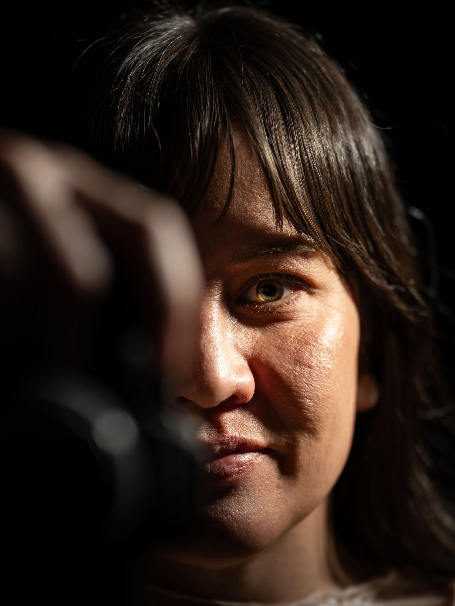 A woman peers through a camera lens in dramatic lighting, with one eye visible and dark background.