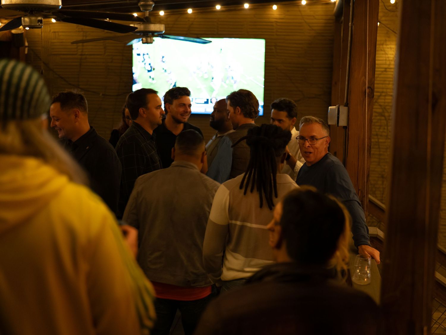 A group of people gathered in a dimly lit room with string lights overhead and a screen visible in the background.