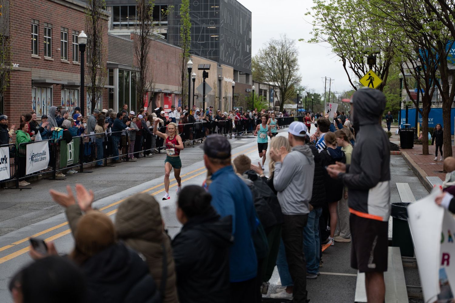 Runners compete in a street race cheered on by spectators lining a barricaded urban course on a cloudy day.