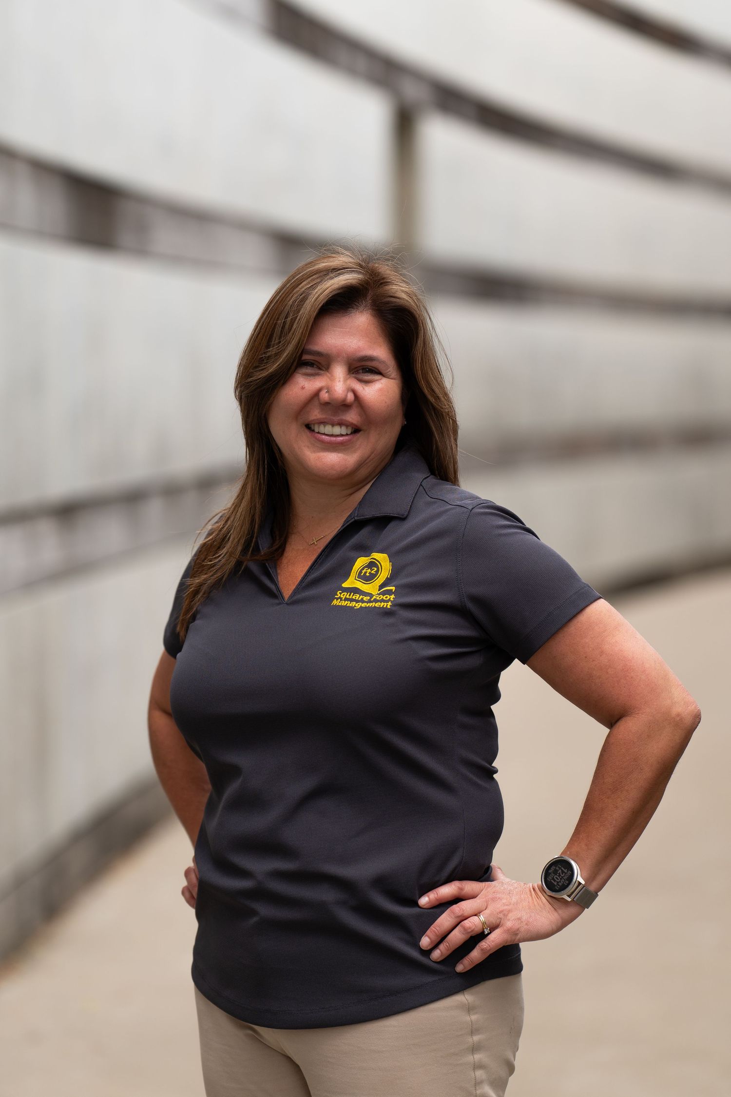 Woman in dark polo shirt with yellow logo smiling confidently, hand on hip, in an outdoor corridor.