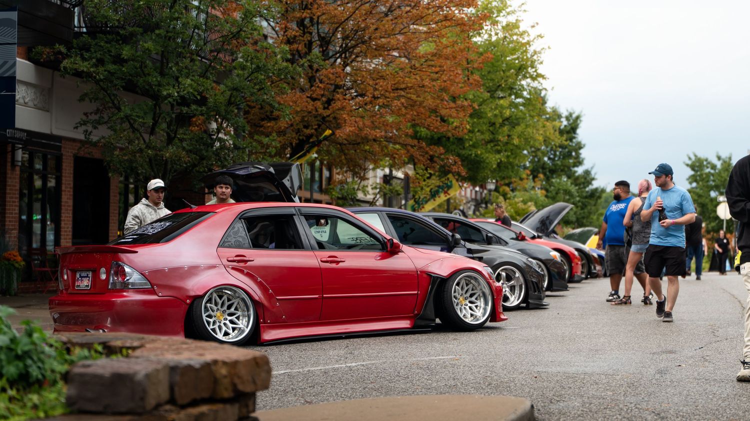Panoramic view of a car show with modified vehicles lined up along a tree-lined street.