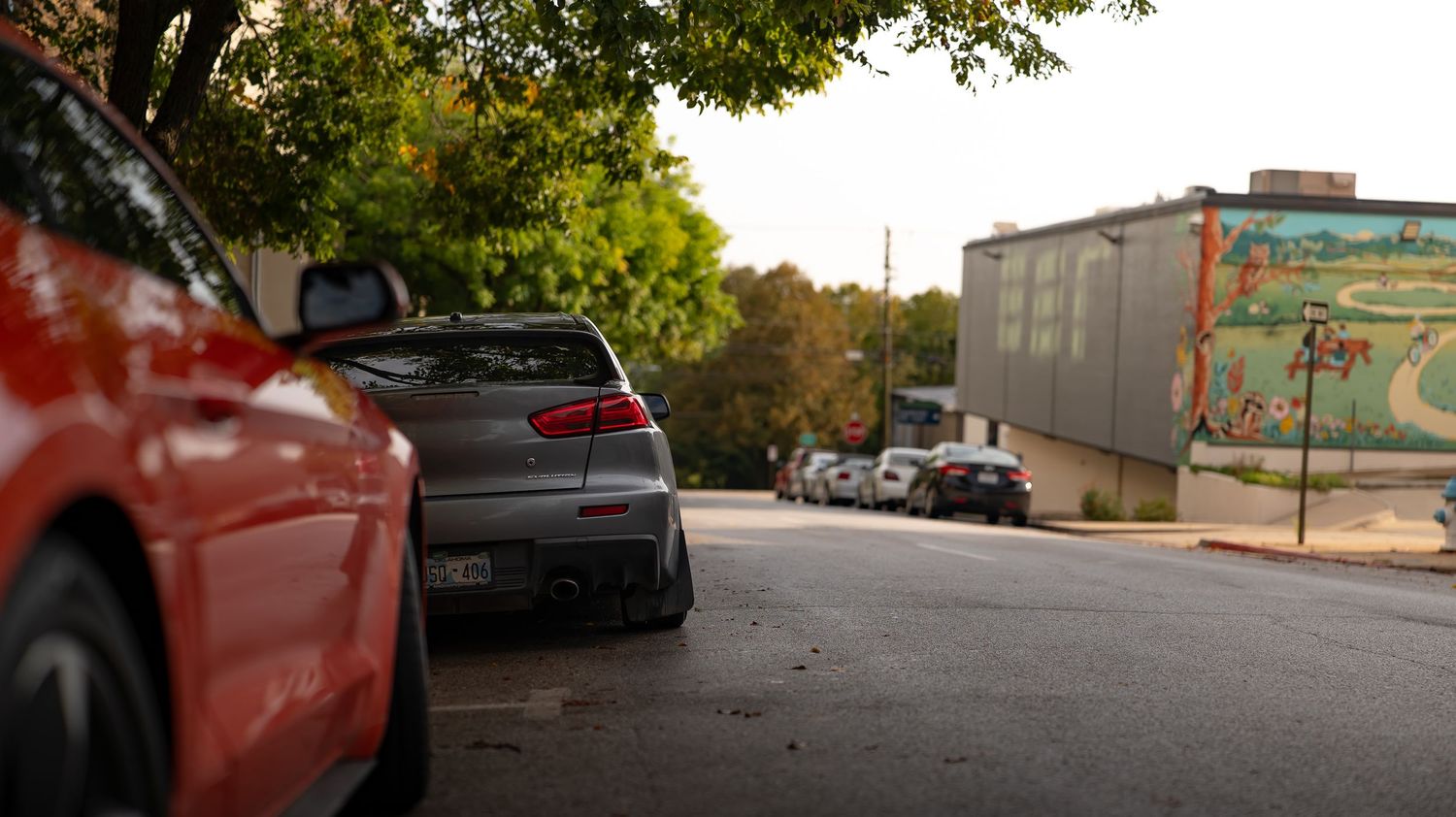 Panoramic view of a tree-lined street with parked cars and brick buildings on a sunny day.