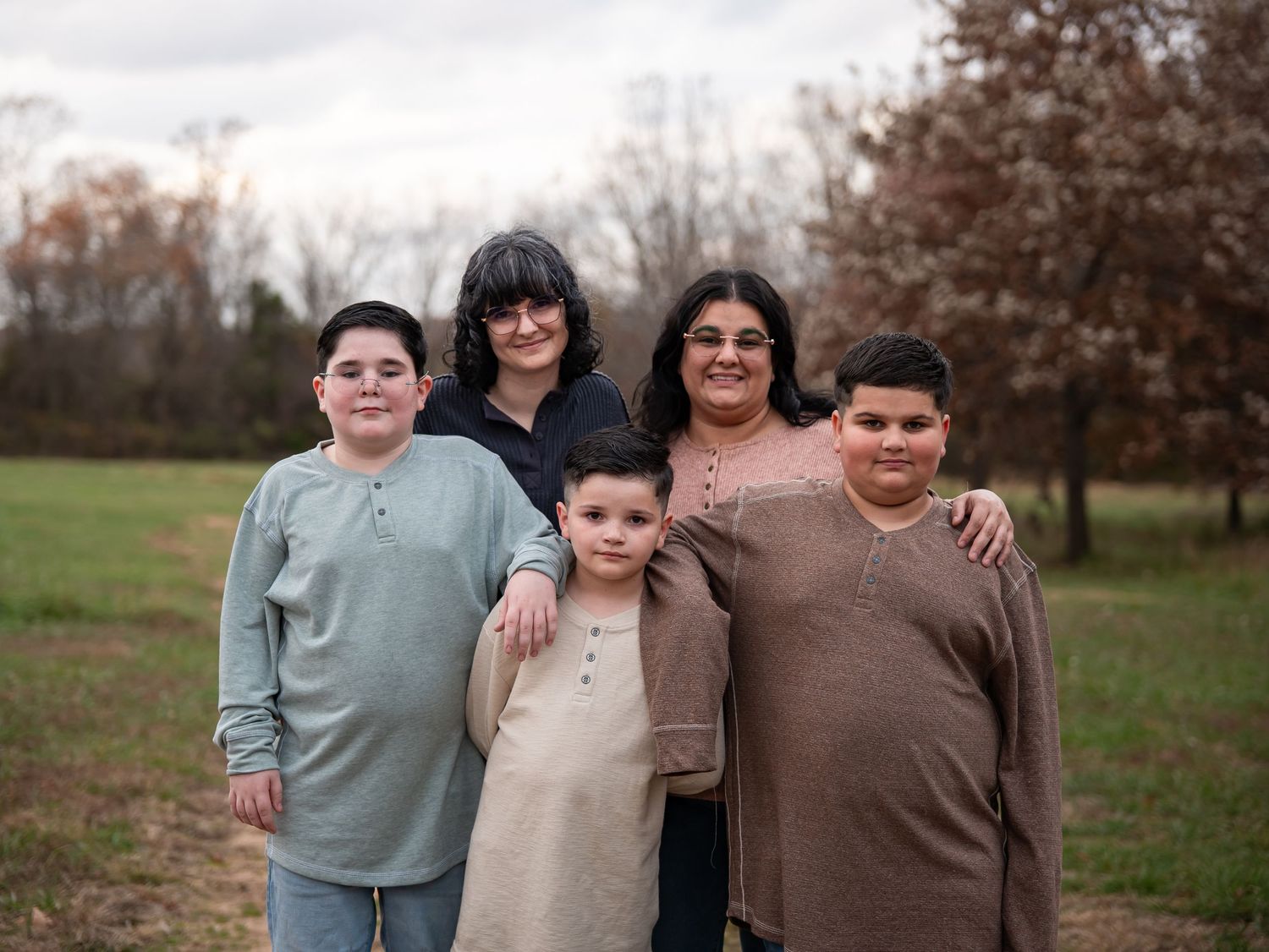 A family group poses together outdoors in casual clothing against an autumn landscape background.