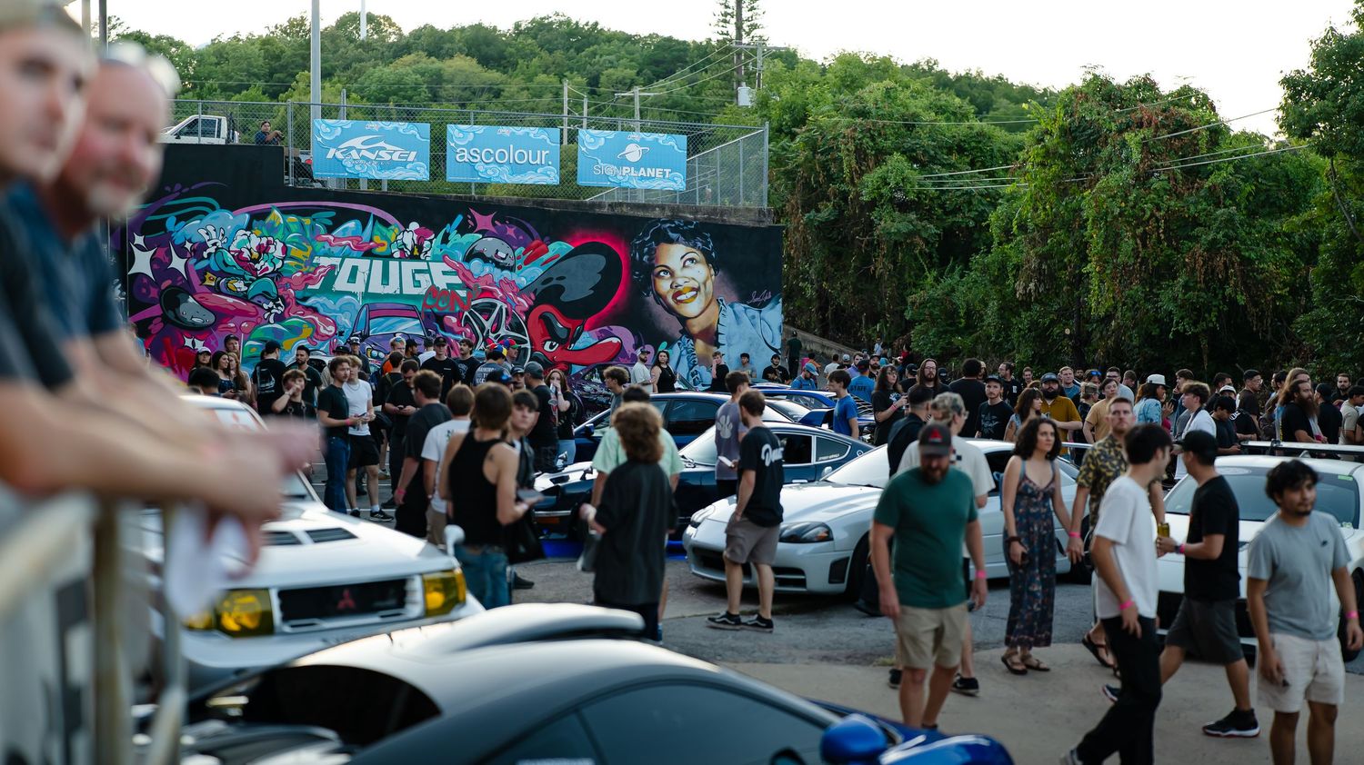 Wide panoramic view of a car meet with vehicles and crowds gathered in a parking lot.