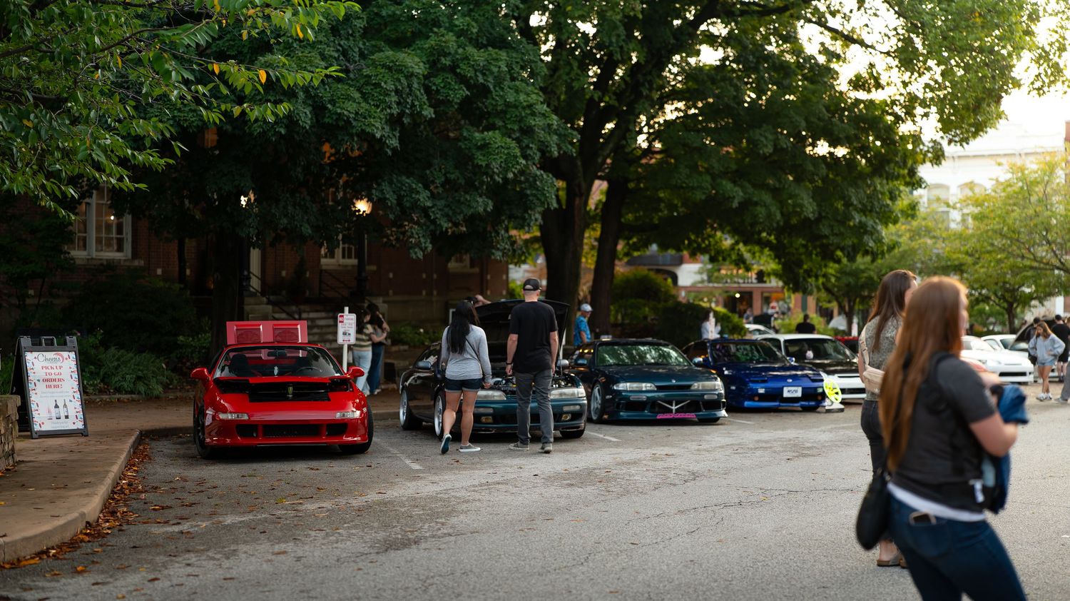 Panoramic view of a car meet showing multiple modified vehicles under tree cover.