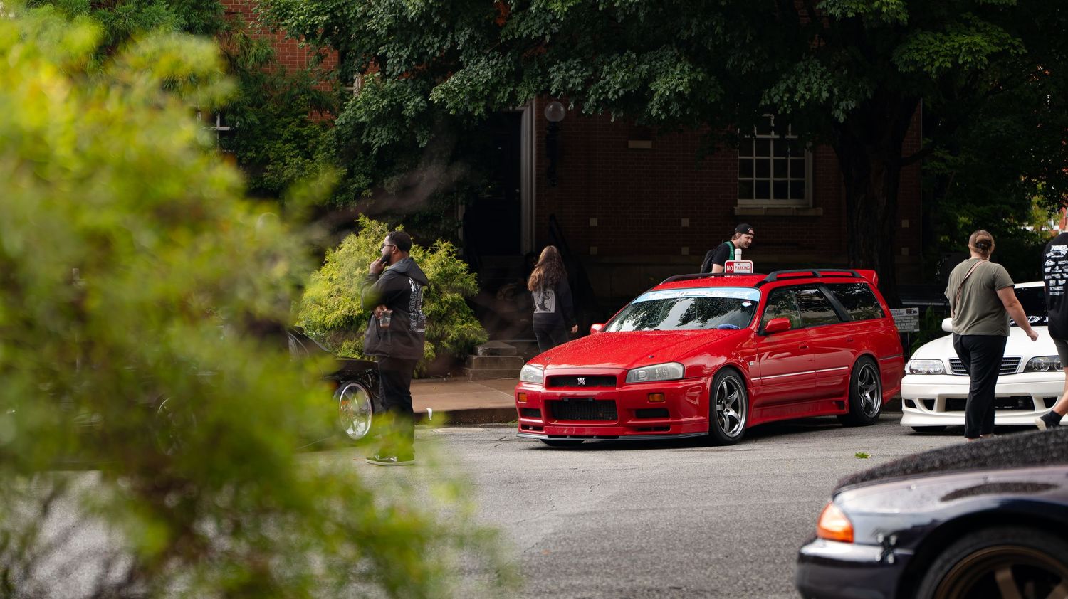 Bright red modified BMW parked under trees at car meet.
