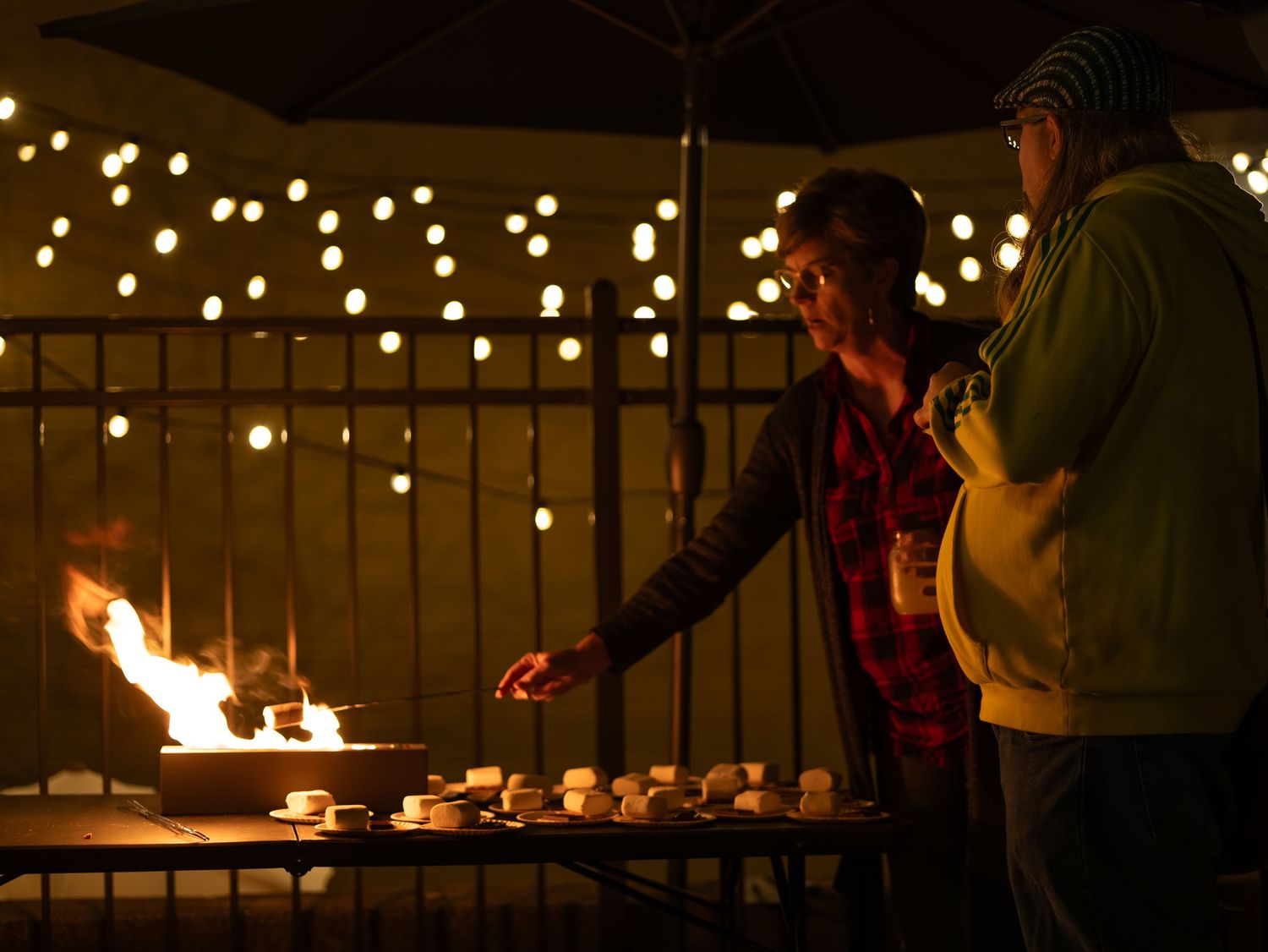 People gather around a fire pit on a deck with string lights overhead during an evening gathering.