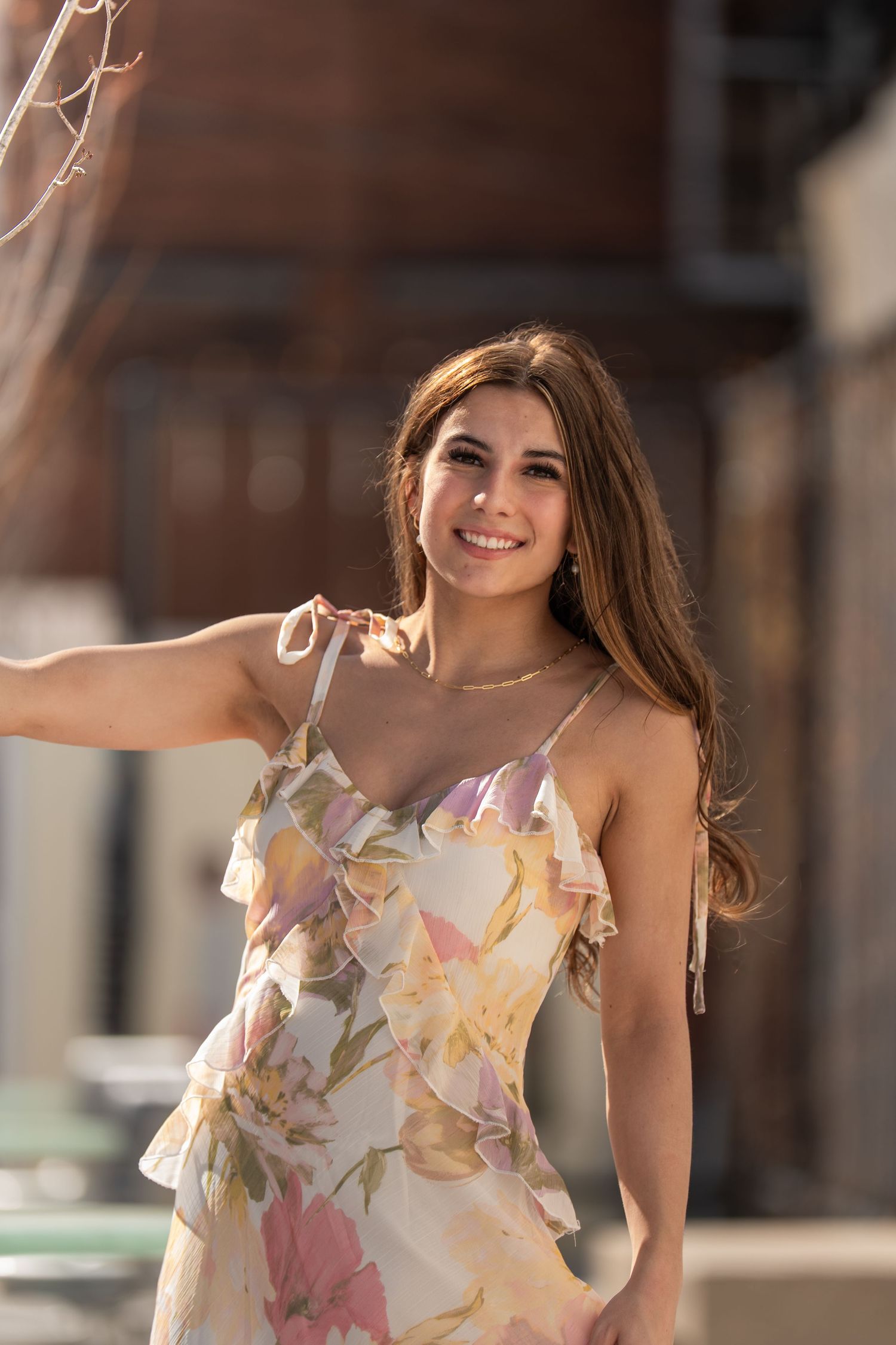Young woman in floral sundress smiling outdoors in warm sunlight, long brown hair, urban background.