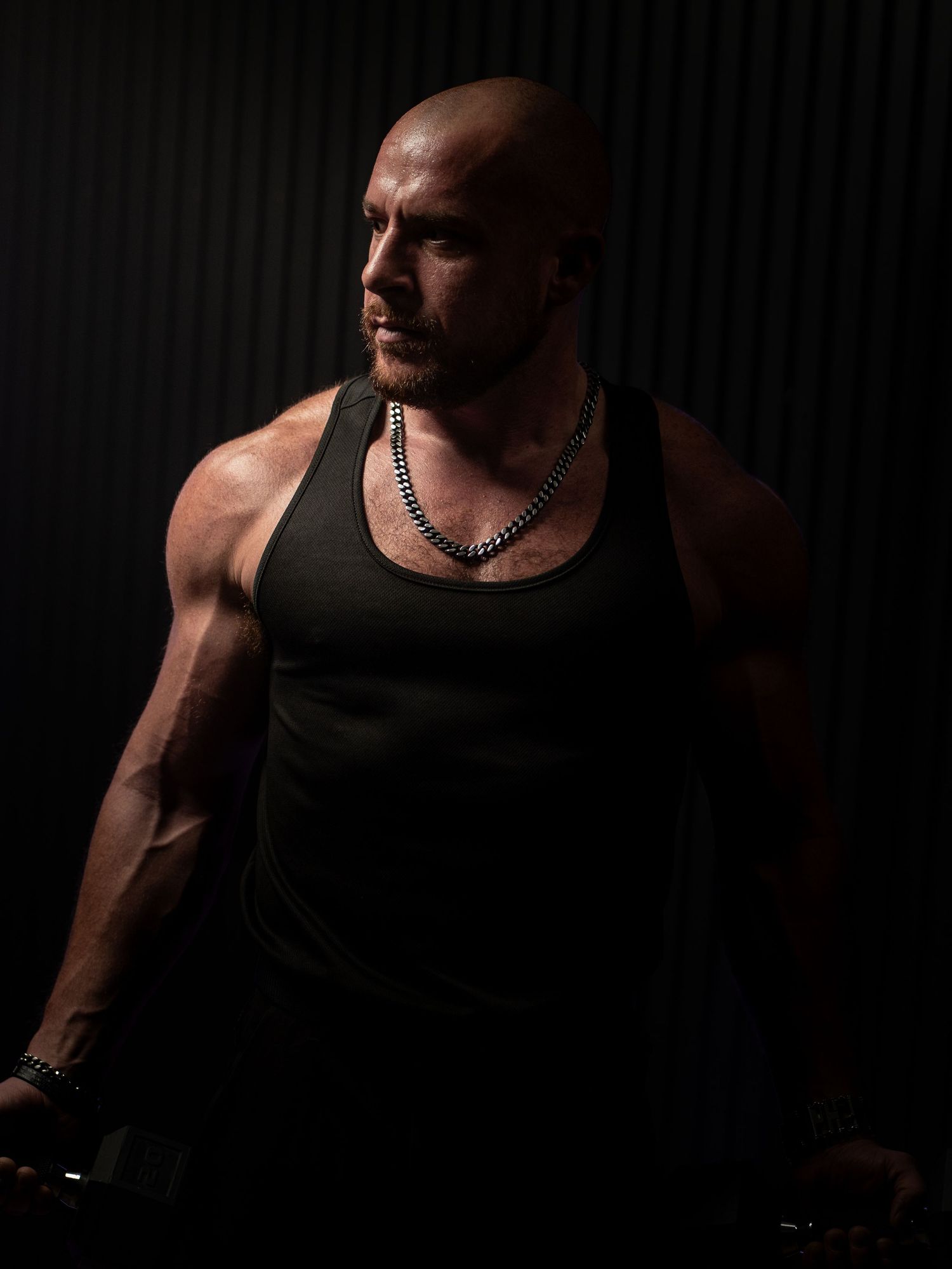 A muscular person in a black tank top poses dramatically against a dark background with moody lighting.