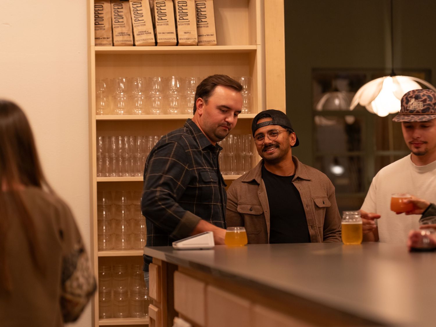 A group of people gathered around a bar counter with wine glasses and shelves of glassware in the background.