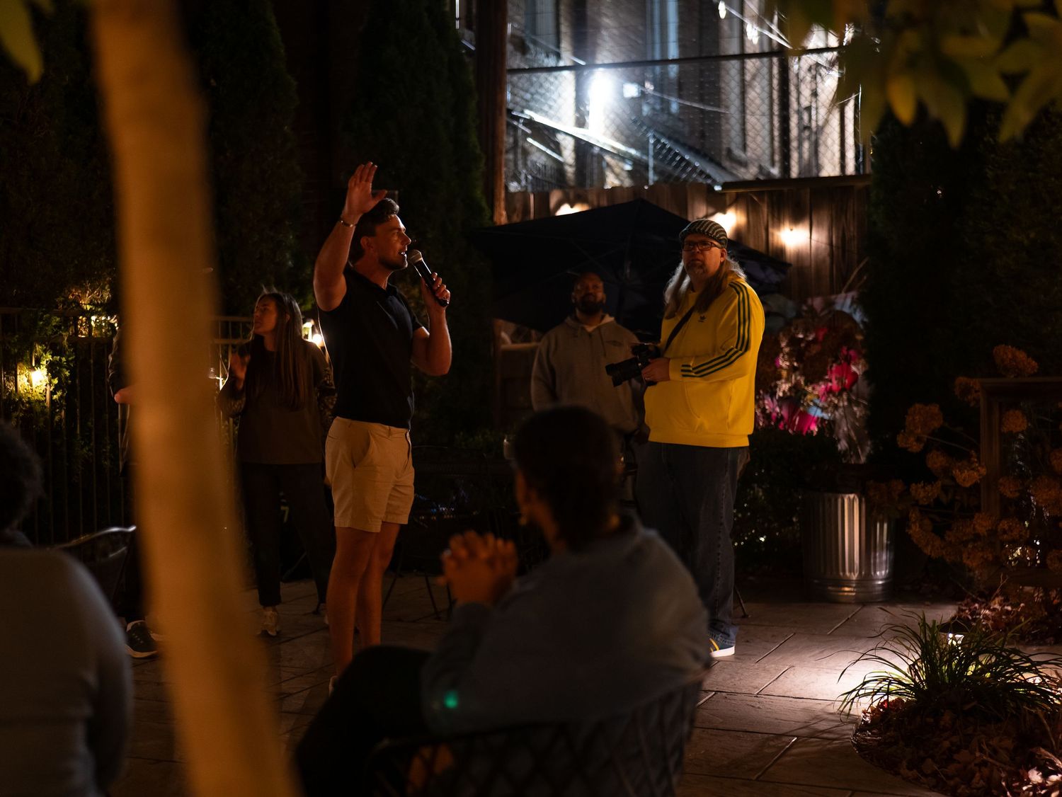 Musicians perform at a dimly lit outdoor venue with string lights and foliage creating an intimate nighttime atmosphere.